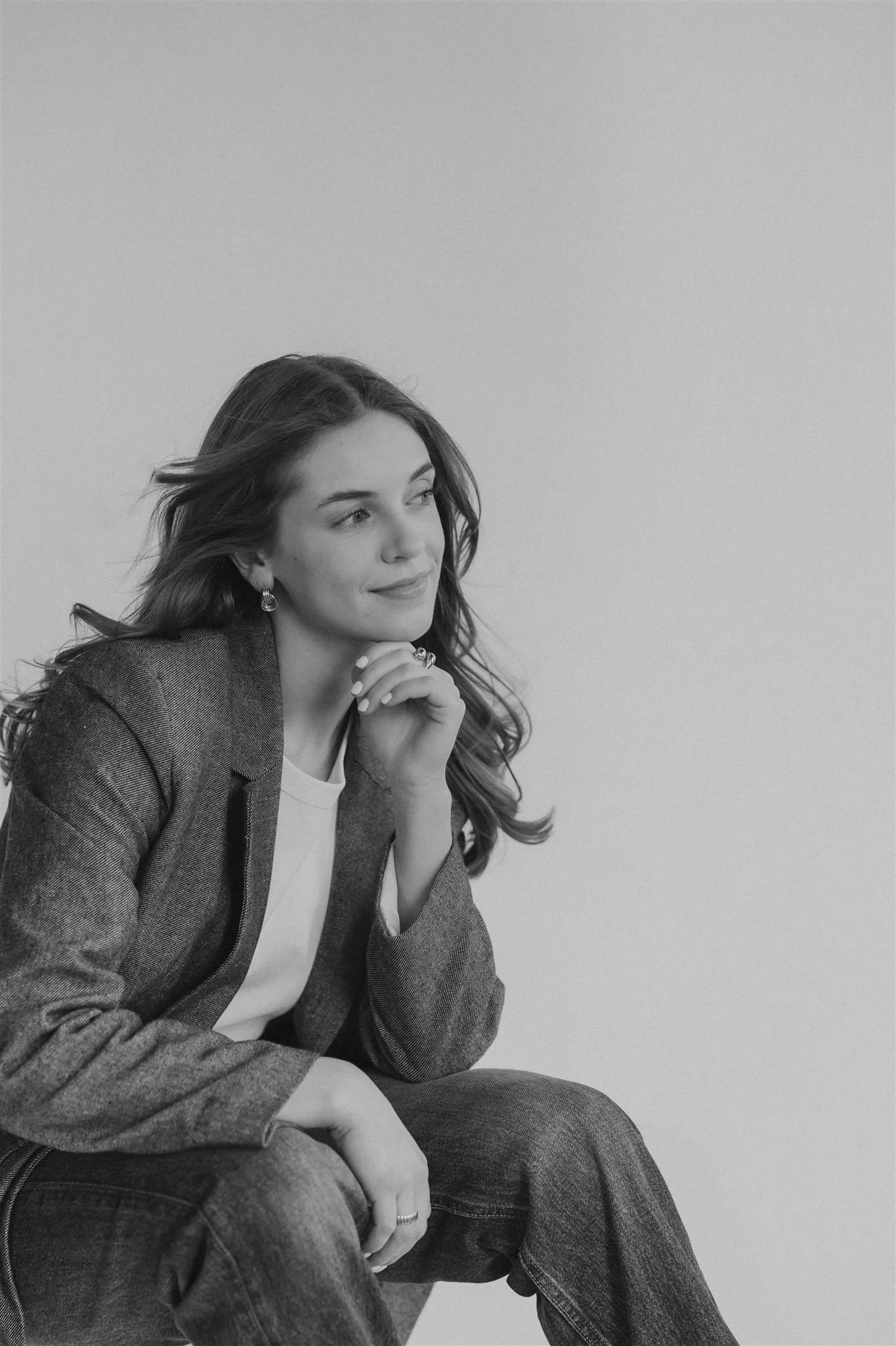 A woman with long wavy hair wearing a blazer, white shirt, and jeans, sitting in a relaxed pose, looking thoughtfully to the side with her hand on her chin.
