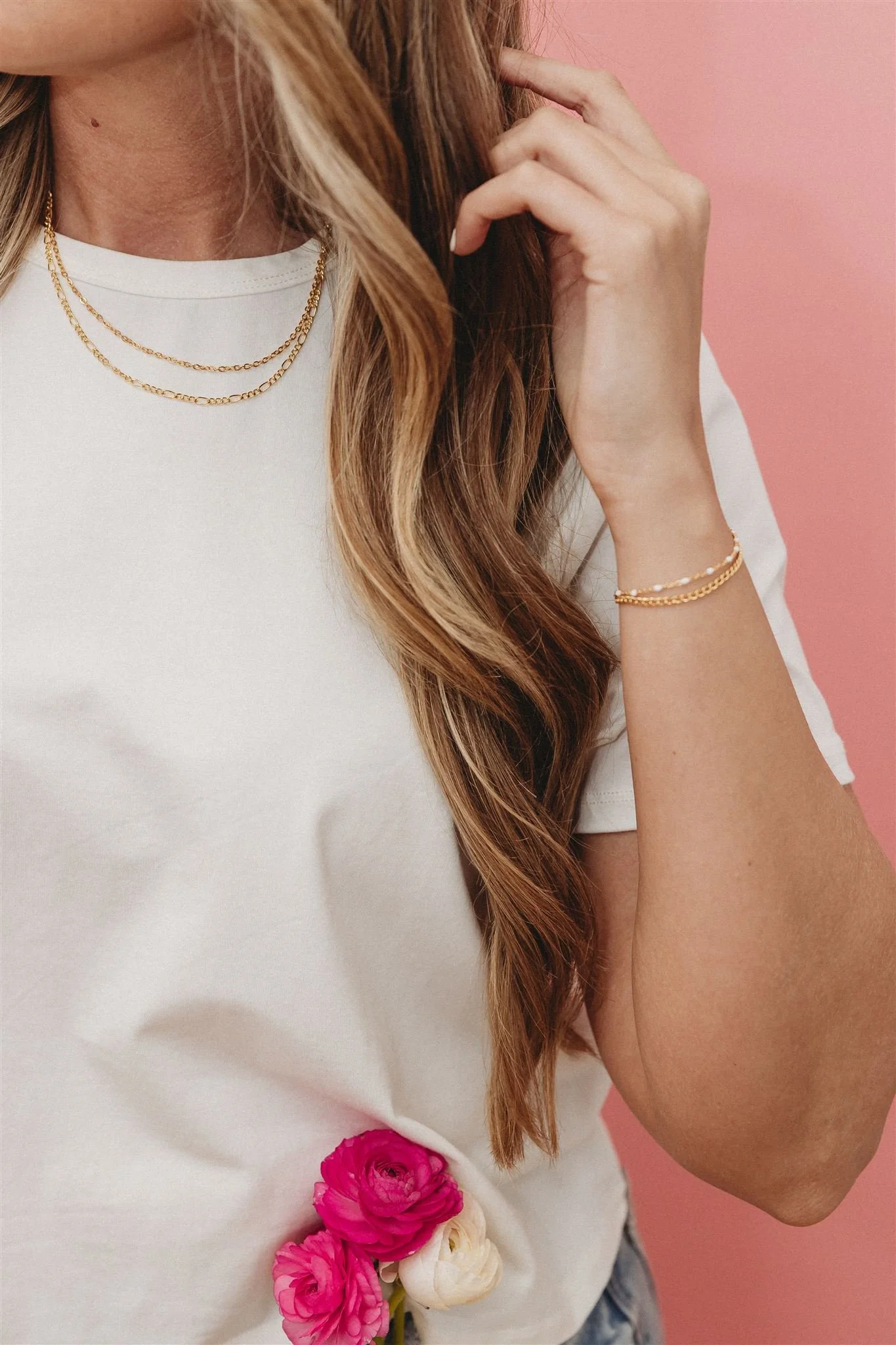 Close-up of a woman wearing layered gold necklaces and a gold bracelet, dressed in a white t-shirt with pink and white flowers tucked in the side, against a pink background.