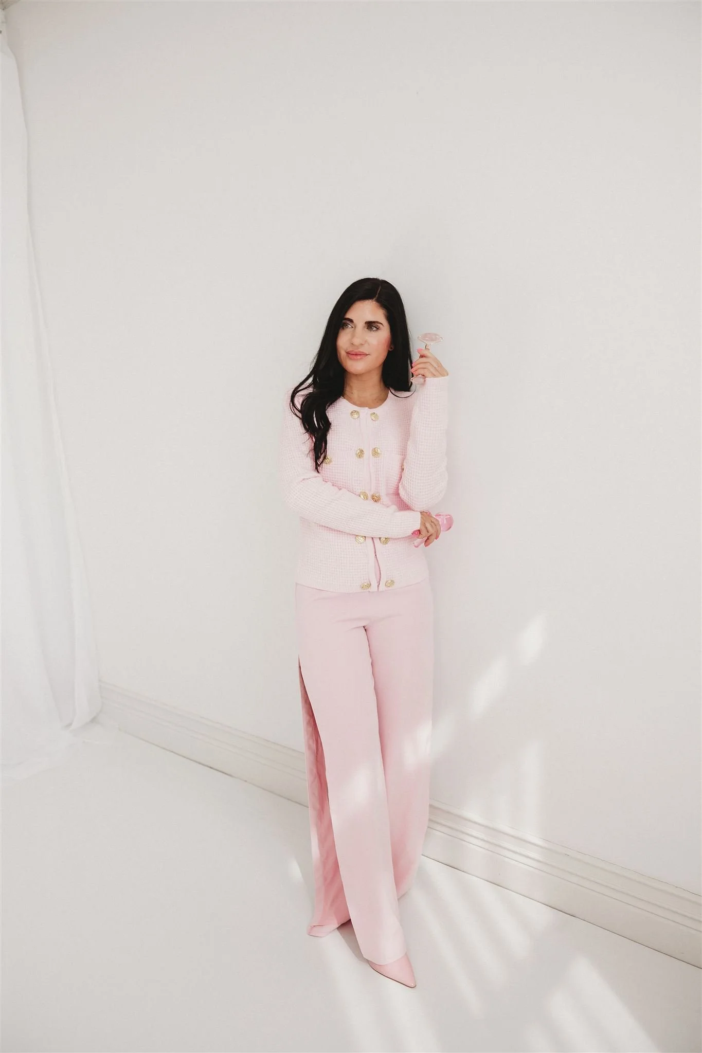 Woman with long dark hair wearing a pink cardigan and pants, holding a pink glass, standing against a white wall.