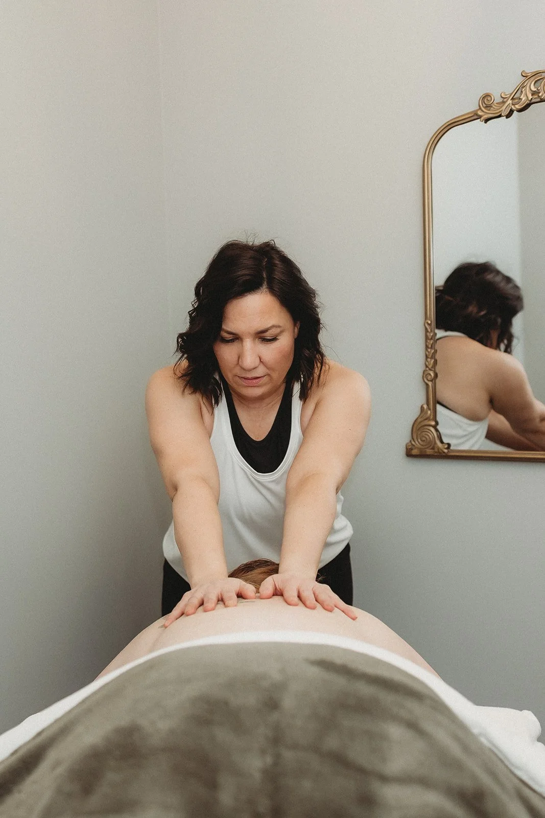 A woman giving a massage to another person lying face down on a massage table in a room with a mirror on the wall.