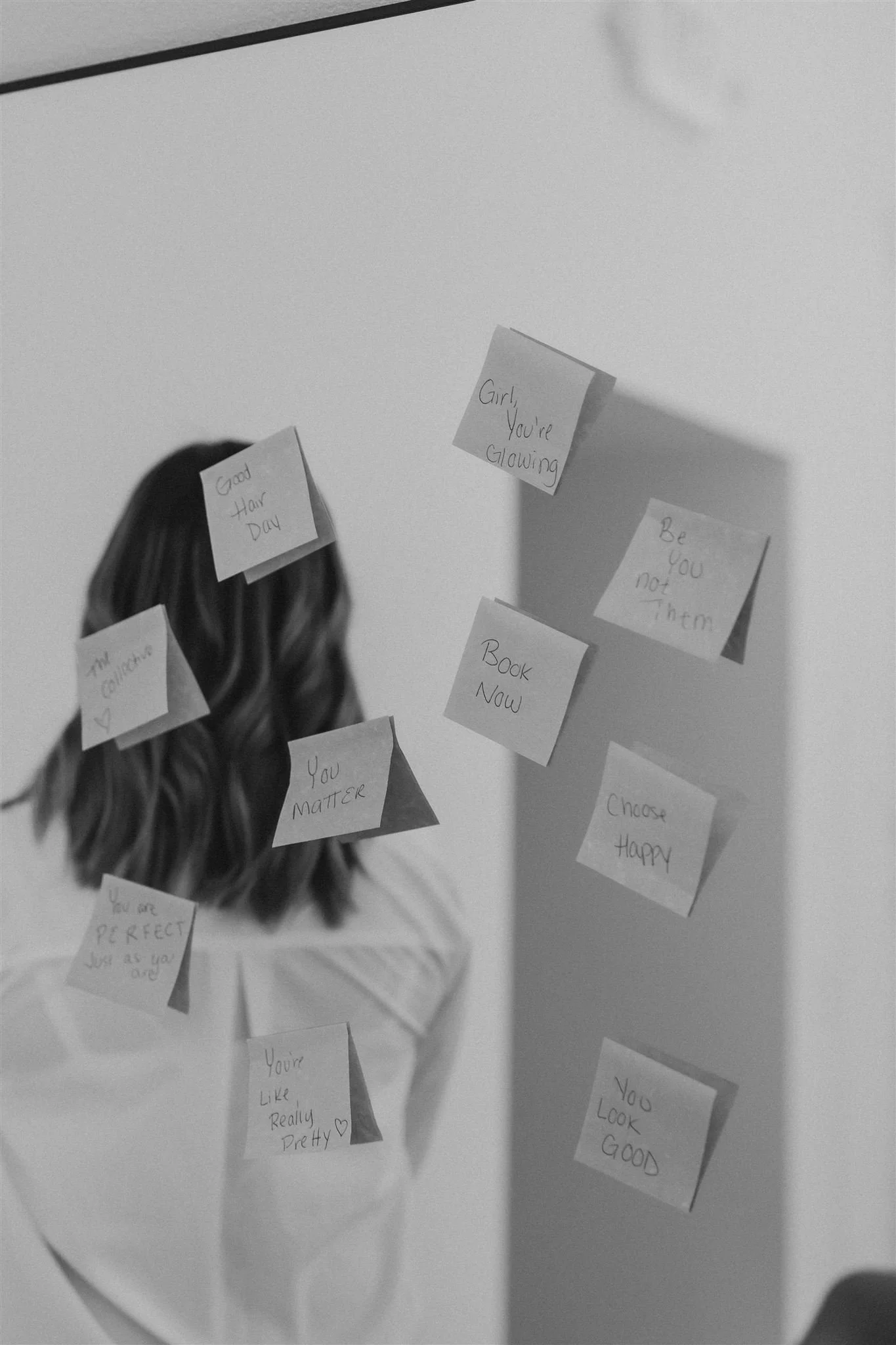 A person with wavy hair looking into a mirror decorated with multiple handwritten notes with positive messages, arranged on the mirror and the person’s back.