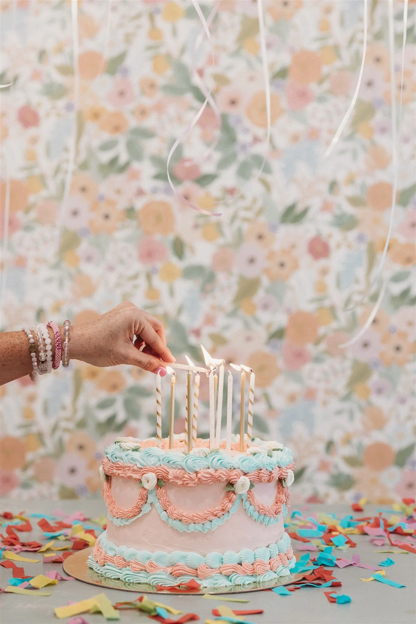 A person is lighting birthday candles on a pink, blue, and white decorated cake with rainbow-colored confetti on a table.