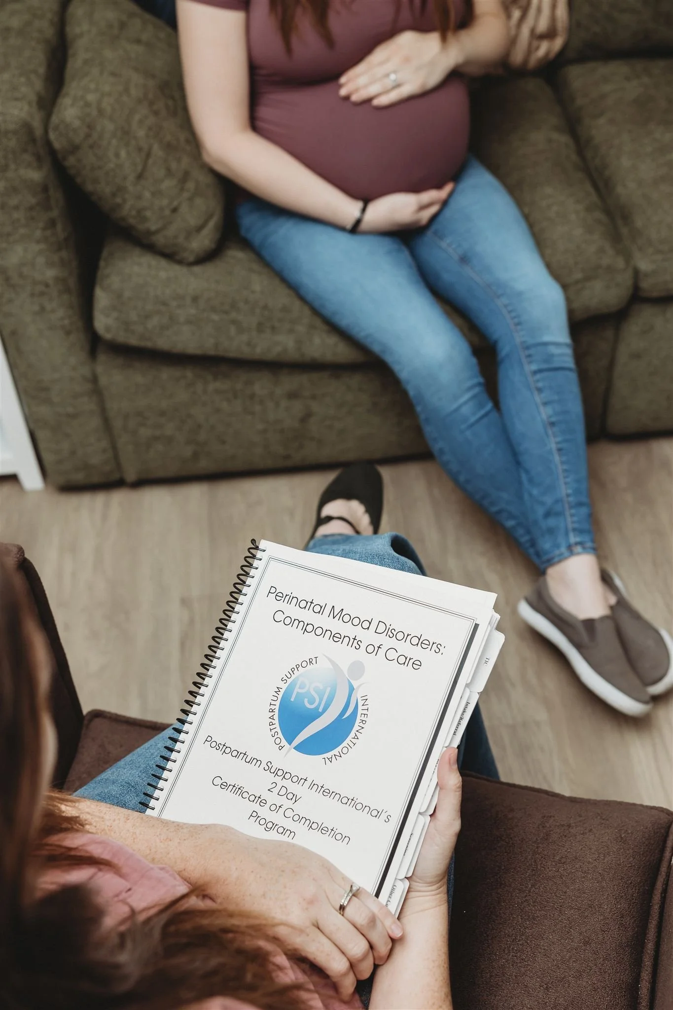 A woman sitting on a brown chair holding a booklet titled 'Perinatal Mood Disorders: Components of Care' with a logo for postpartum support international. In front of her, another person is sitting on a brown couch, wearing blue jeans and a maroon shirt, with hands resting on her pregnant belly. The scene appears to be a counseling or support session.
