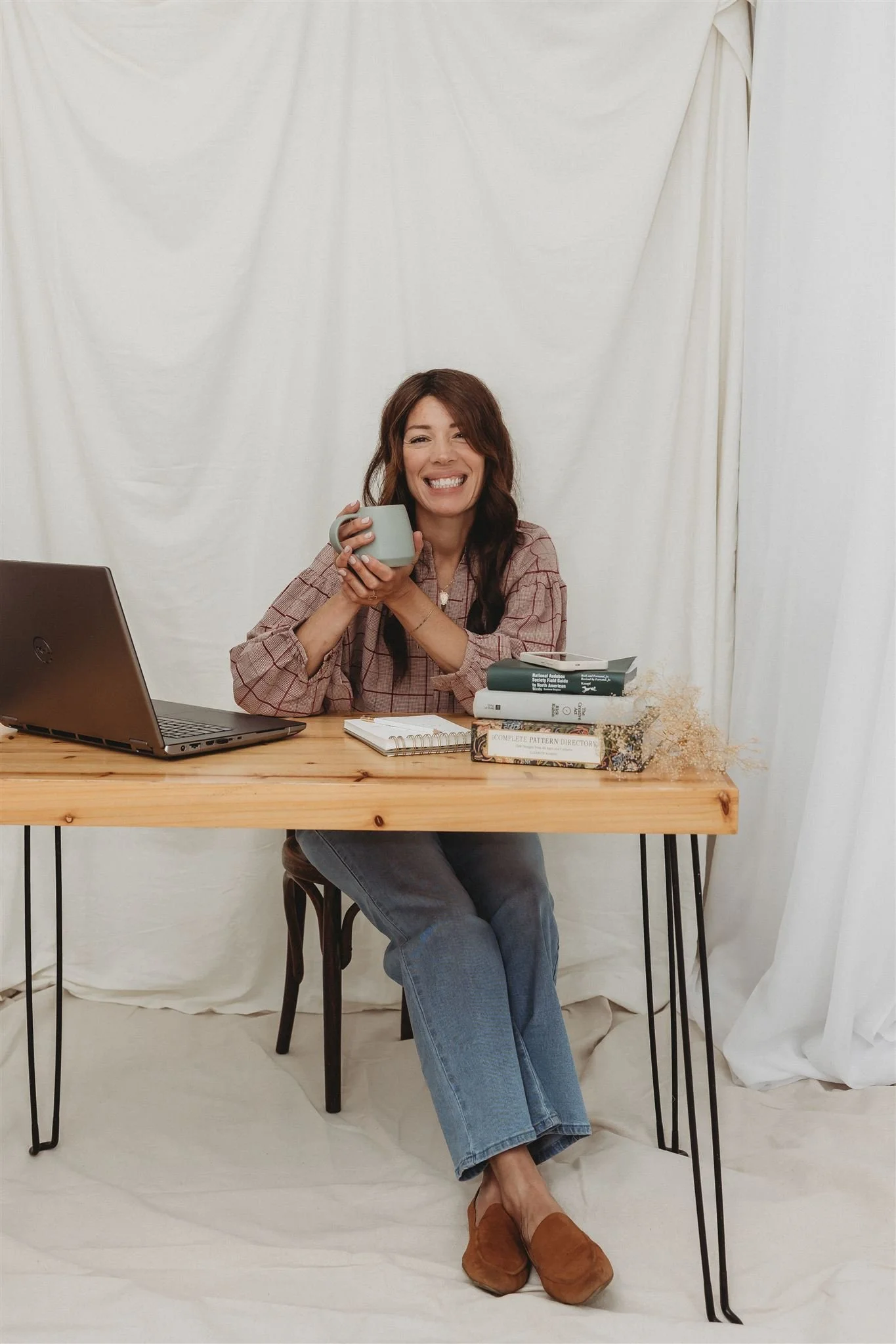 A woman smiling, sitting at a wooden desk with a laptop, books, and a notebook, holding a mug, in front of a white curtain backdrop.