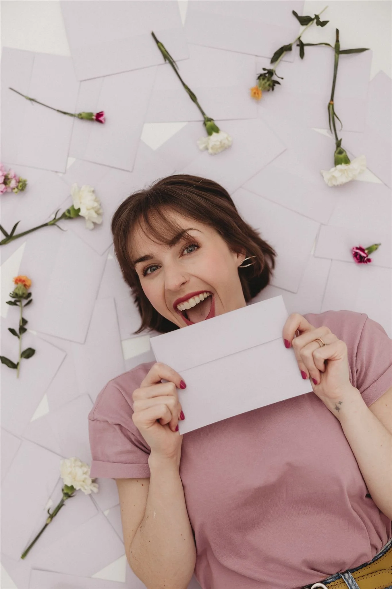 A woman lying on a floor surrounded by scattered white envelopes and colorful flowers, holding a white paper folder and smiling.