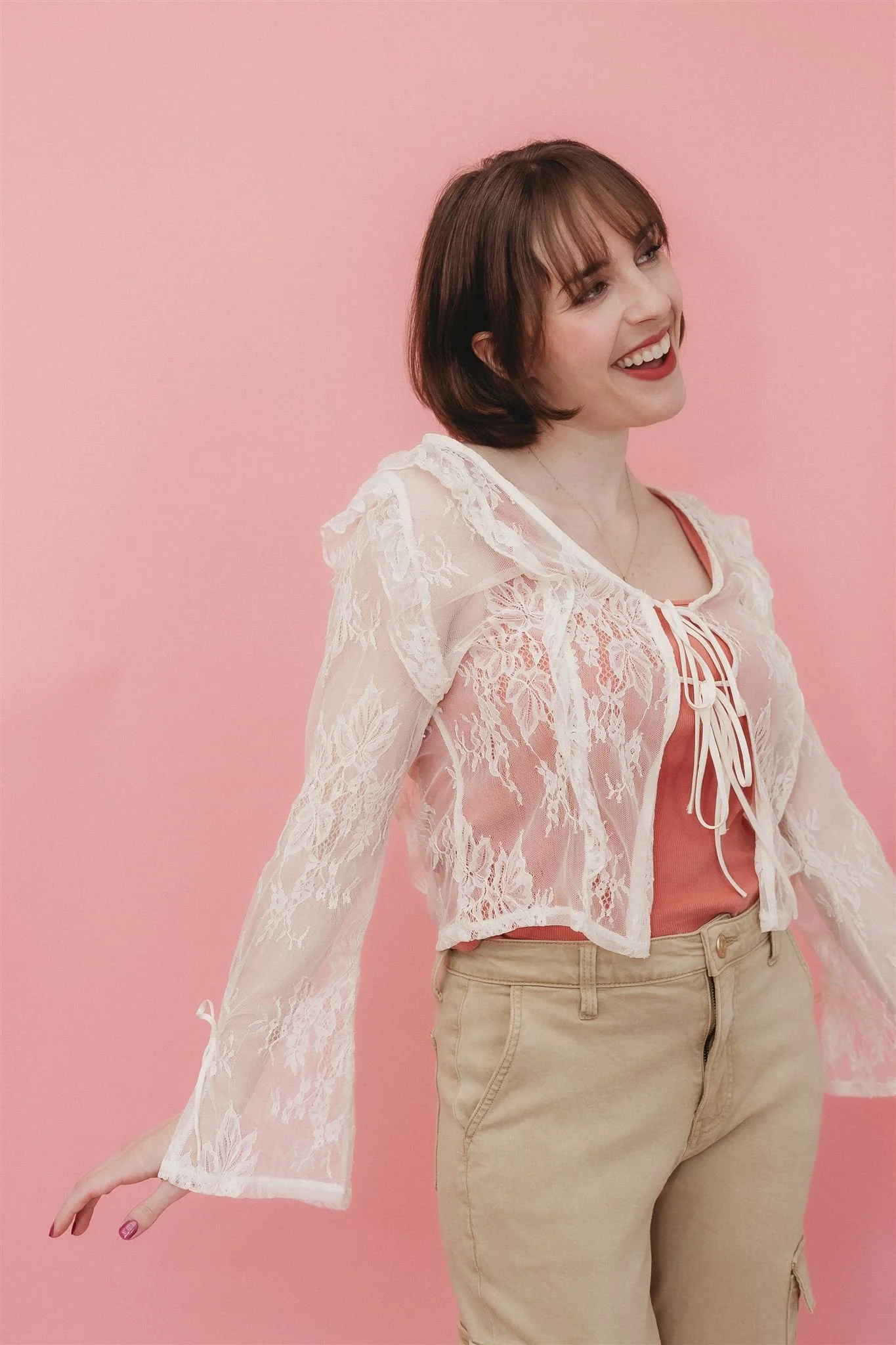 A young woman with short brown hair, smiling, wearing a white lace jacket over a pink top, beige pants, standing against a pink background.