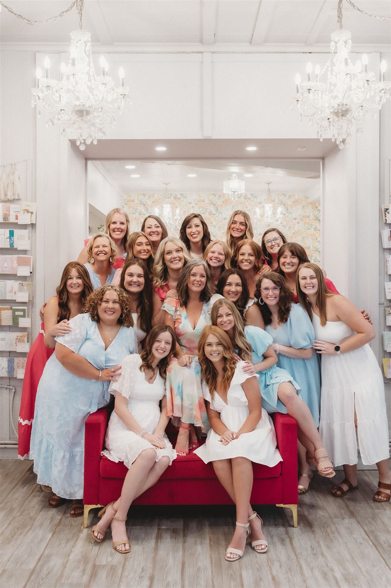 A large group of women smiling for a photo inside a brightly decorated room with chandeliers and floral wallpaper, some wearing dresses in pastel colors.