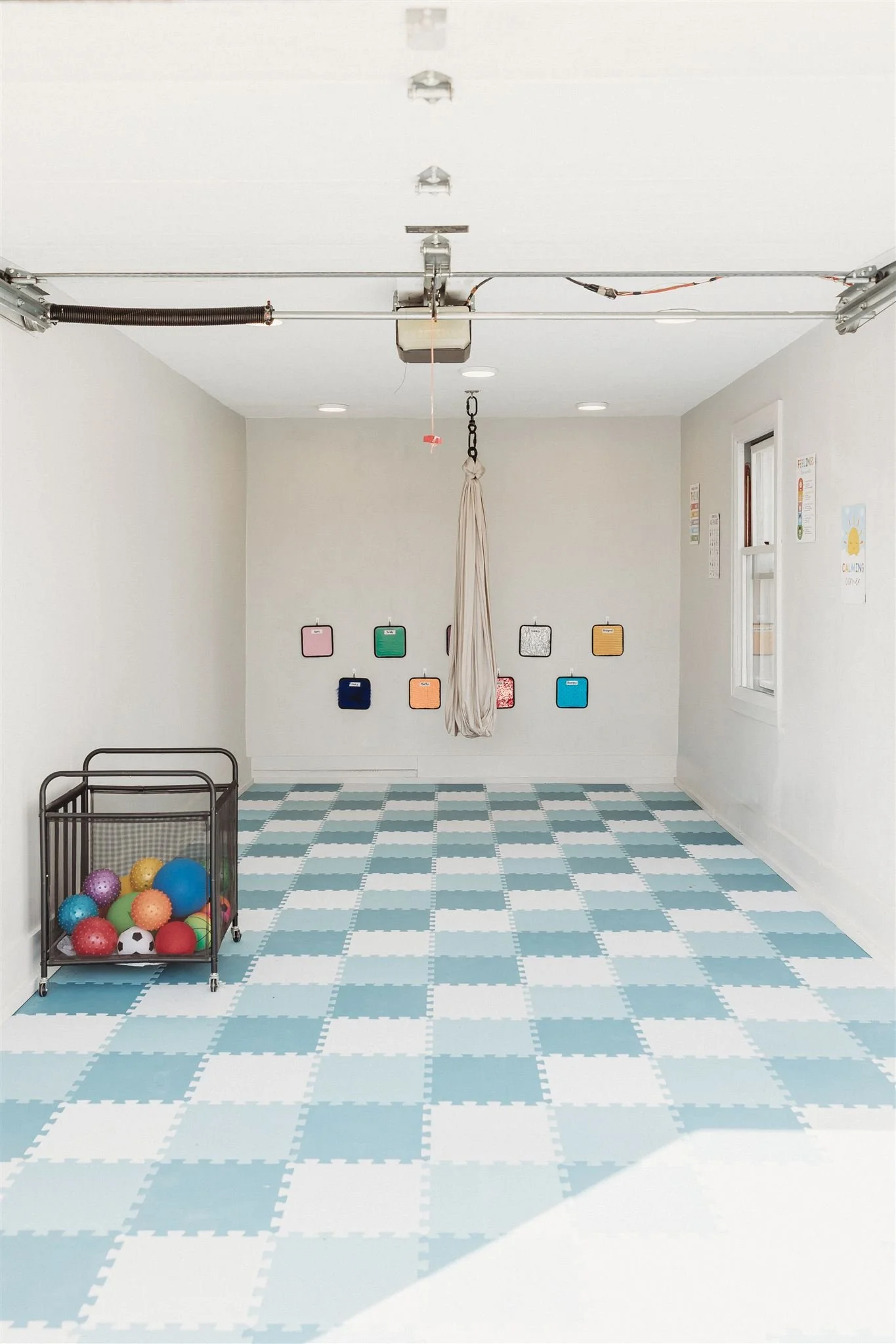 Empty sensory room with a checkered blue and white foam floor, a collapsible cart filled with colorful textured balls, a white wall with small mounted colorful square cushions, and a white ceiling with garage door mechanism and a fabric hanging from the ceiling.