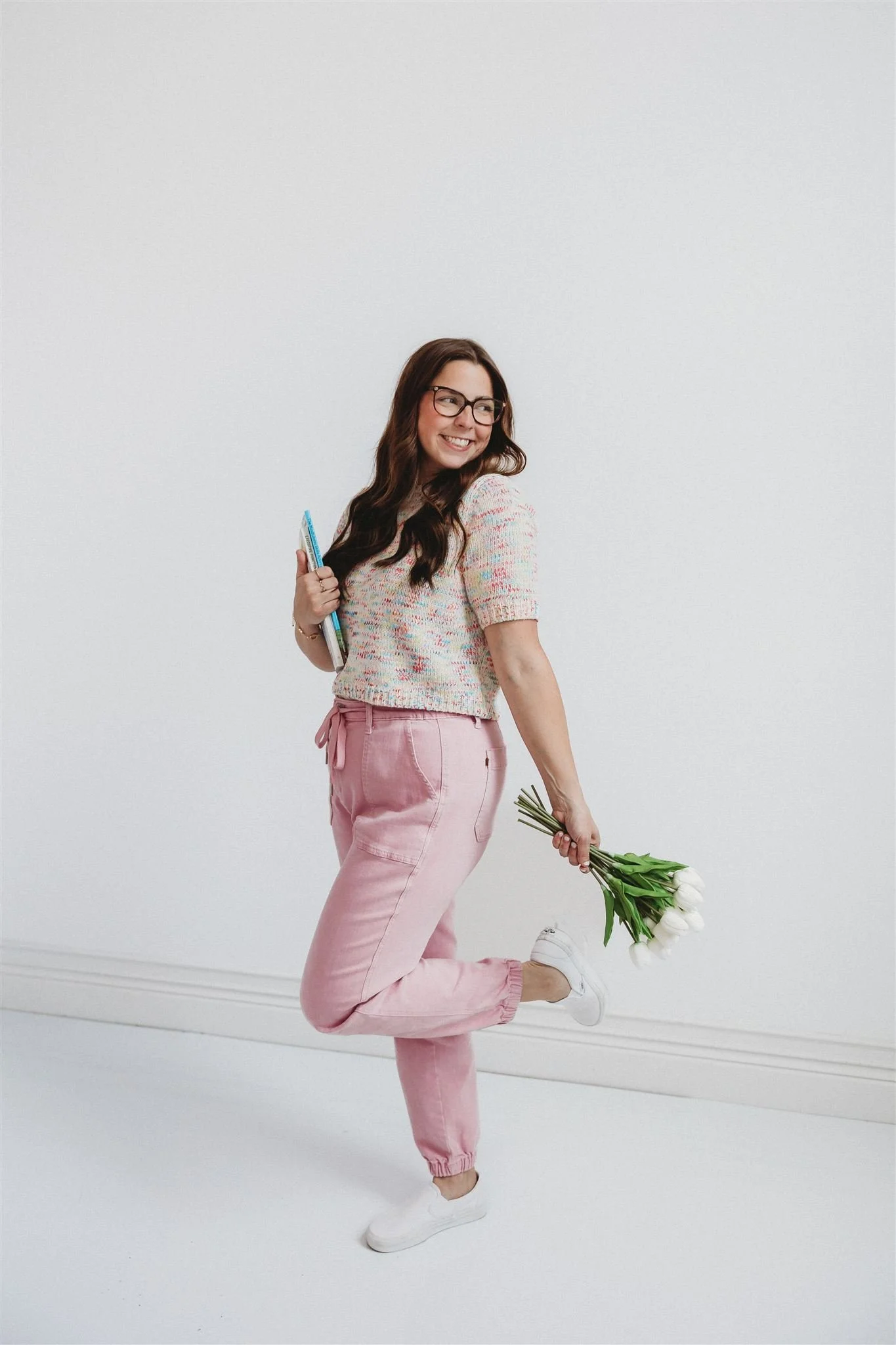 A smiling woman with long brown hair and glasses poses playfully against a white wall, wearing a multicolored knit top and pink jogger pants with white slip-on sneakers, holding a book under one arm and a bunch of white tulips in her other hand.