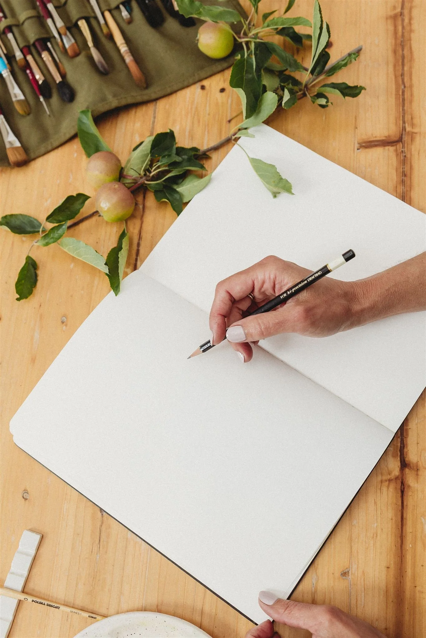 A person holds a black pencil with a gold tip over an open, blank white notebook on a wooden table. On the table, there are green leafy branches with small round fruits, possibly apples, and a set of paintbrushes in a green fabric roll.