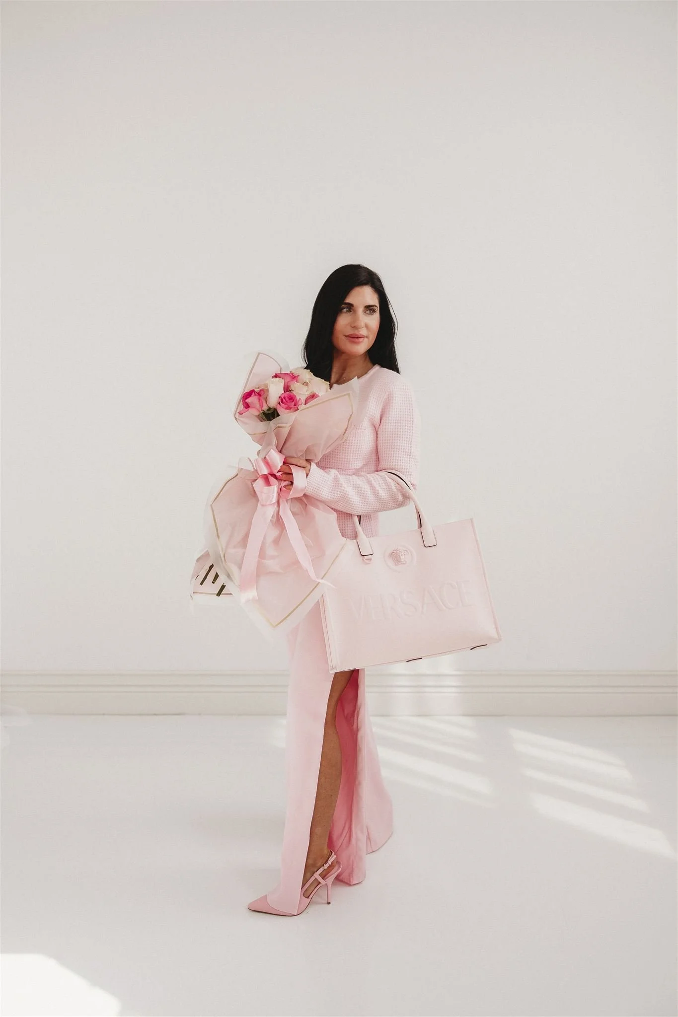 A woman dressed in a pink outfit holding a bouquet of pink roses and a large pink Versace shopping bag, posing in a minimalistic white room.
