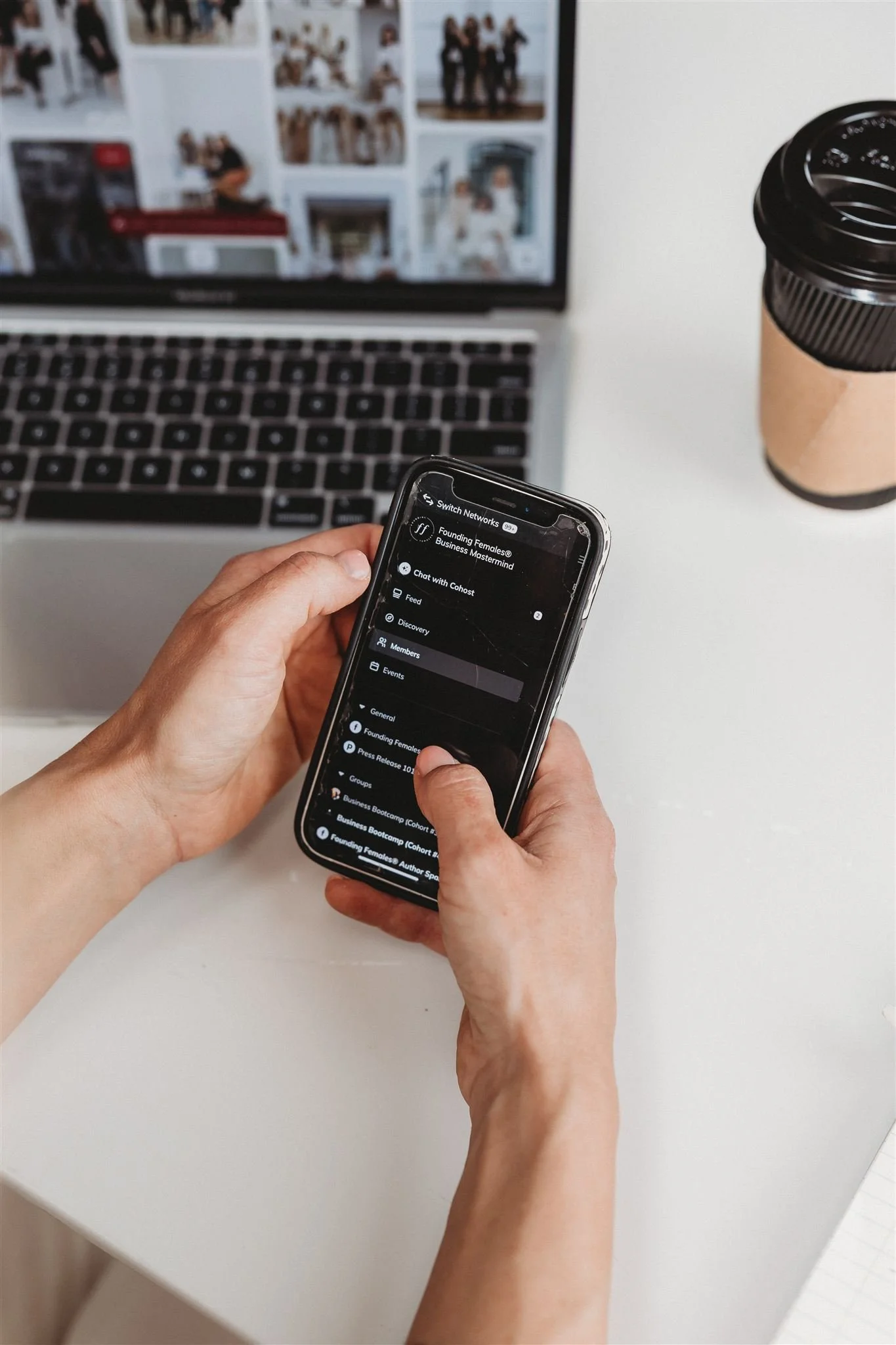 Person holding a smartphone at a white desk with a laptop and a coffee cup, browsing a social media app.