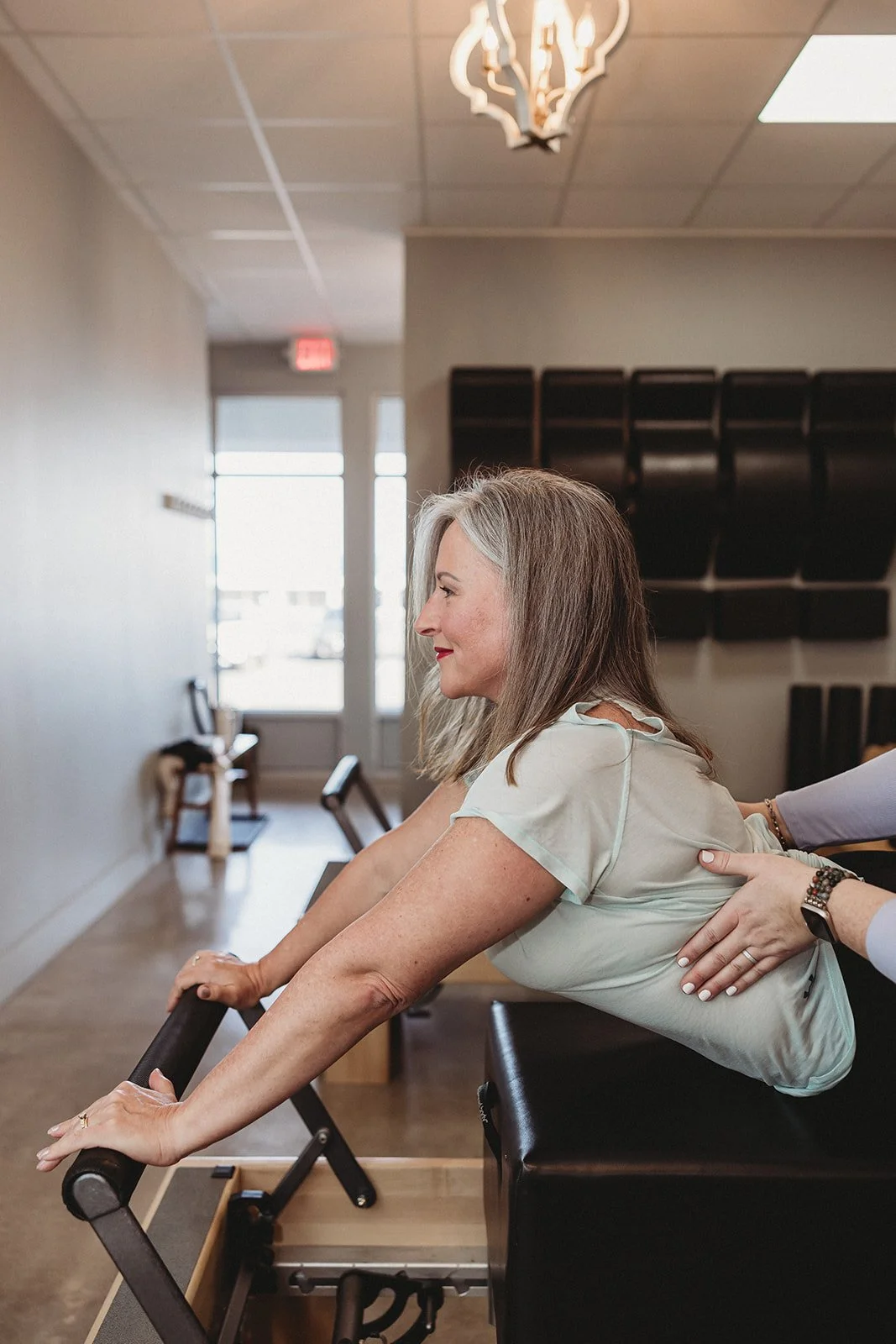A woman with gray hair and a beige top performs a backbend on a Pilates reformer machine in a fitness studio, with a gentle smile and a supportive hand on her back.
