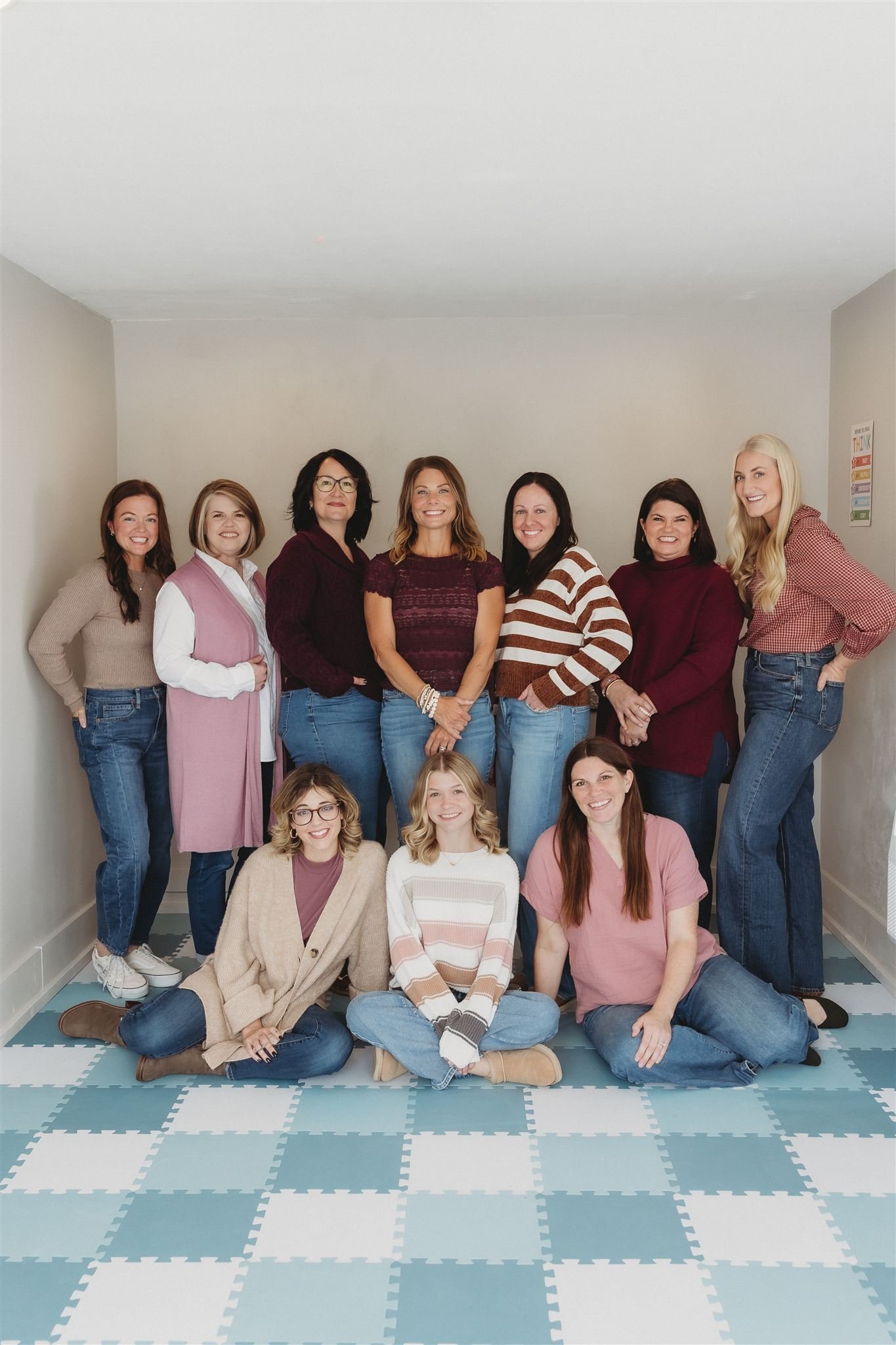 A group of eleven women posing in a room with light-colored walls and a checkered floor, smiling and looking at the camera.