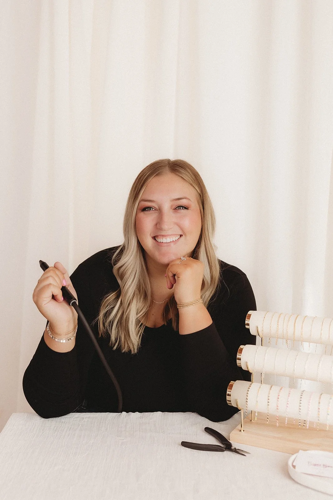 A smiling woman with blonde hair, dressed in black, sitting at a table with jewelry display stands, jewelry pliers, and jewelry she is working on.