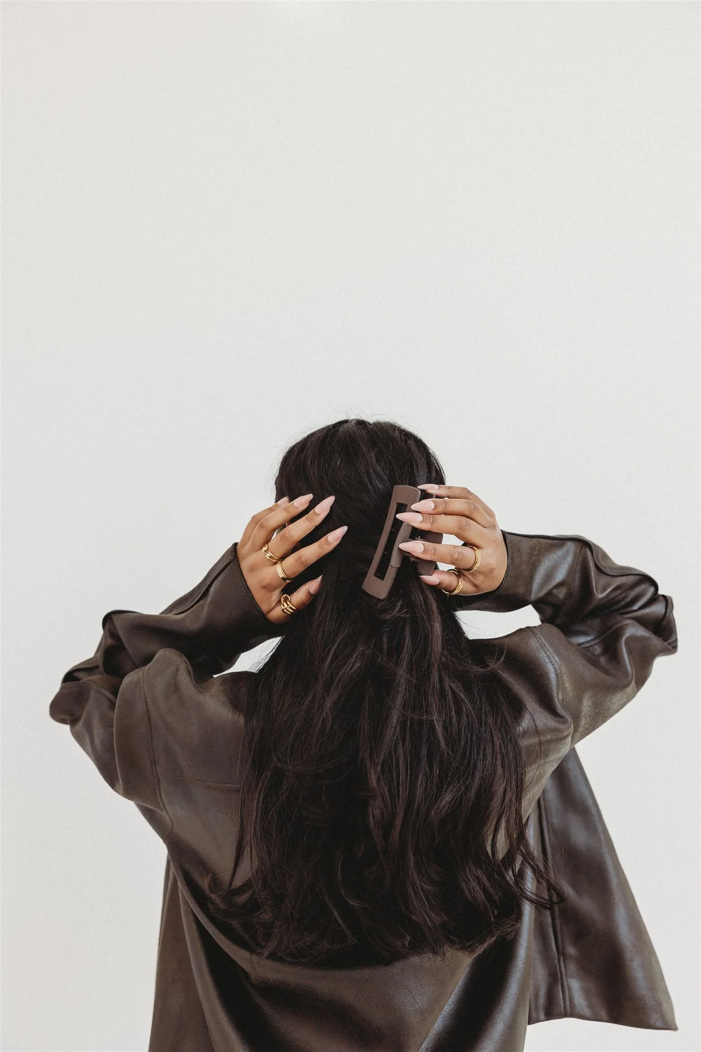 A woman with long, dark hair, wearing multiple rings and a brown leather jacket, is fixing her hair with a hair clip against a plain white background.
