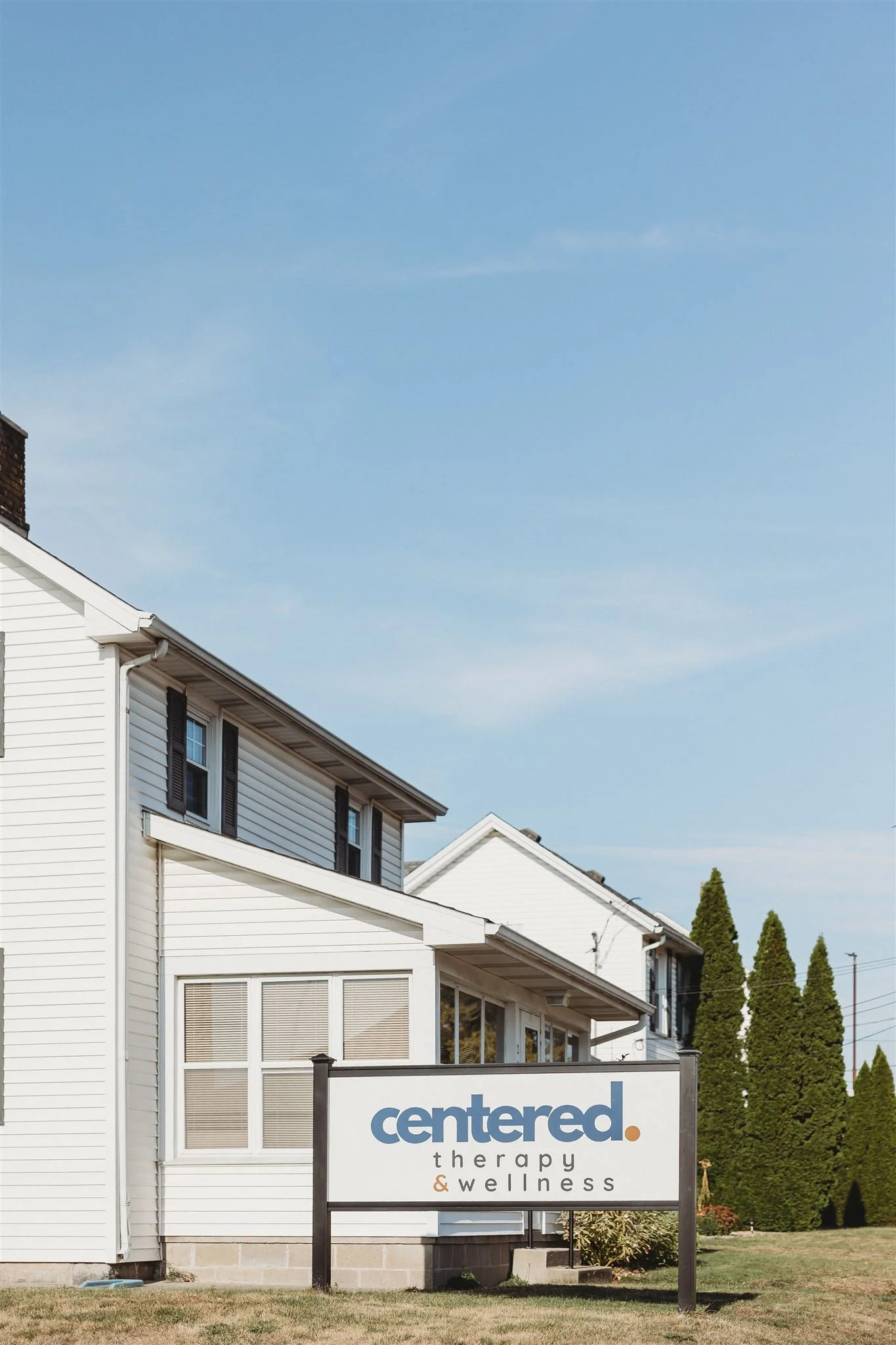 A sign in front of a white building reads 'centered. therapy & wellness'. The building has multiple windows and is surrounded by a grassy area with tall evergreen trees. The sky above is clear and blue.
