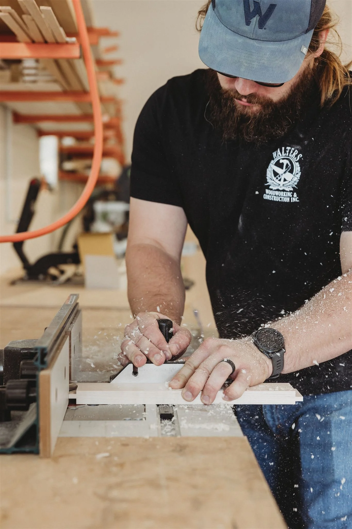 Man woodworking at a workbench in a workshop, wearing a blue cap, glasses, and a black shirt with a woodworking logo, using a hand tool to cut a piece of wood with sawdust around.