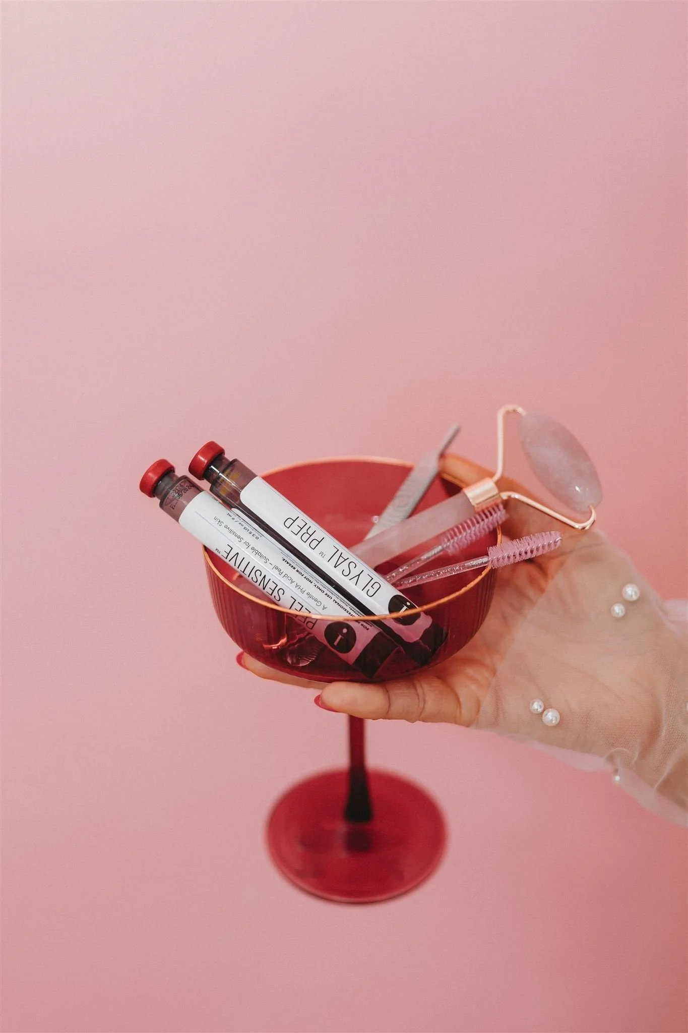 Hand holding a red coupe glass with makeup tools, including two small vials labeled 'GLY-SAL PREP,' a mascara spoolie, eyelash glue, and a jade roller against a pink background.