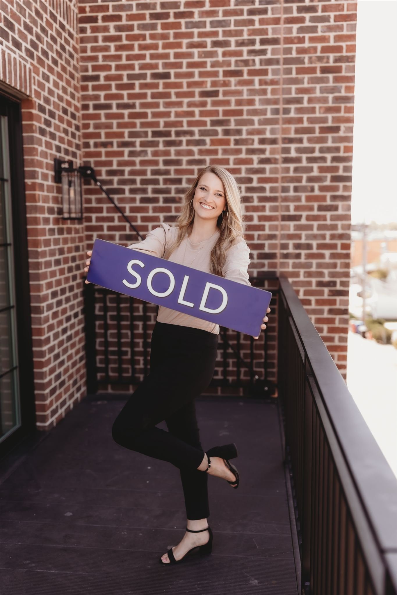 A woman on a balcony holding a purple sign that says 'SOLD', smiling at the camera.