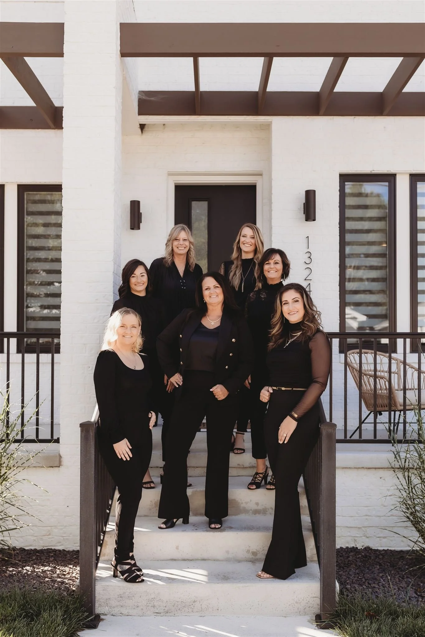 Group of eight women standing on steps outside a modern white brick house, dressed in black.