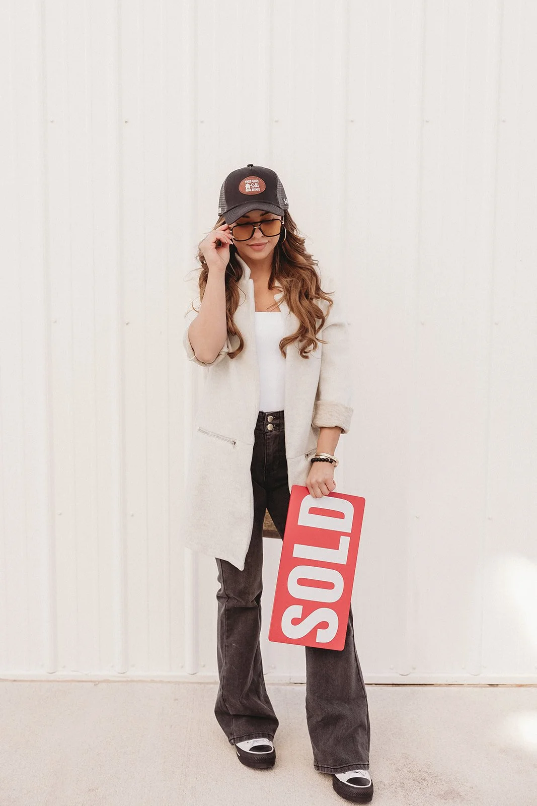 Woman with long curly hair wearing a black cap, glasses, a white shirt, a beige coat, black jeans, and sneakers, holding a red sign that says 'SOLD', standing against a plain white wall.