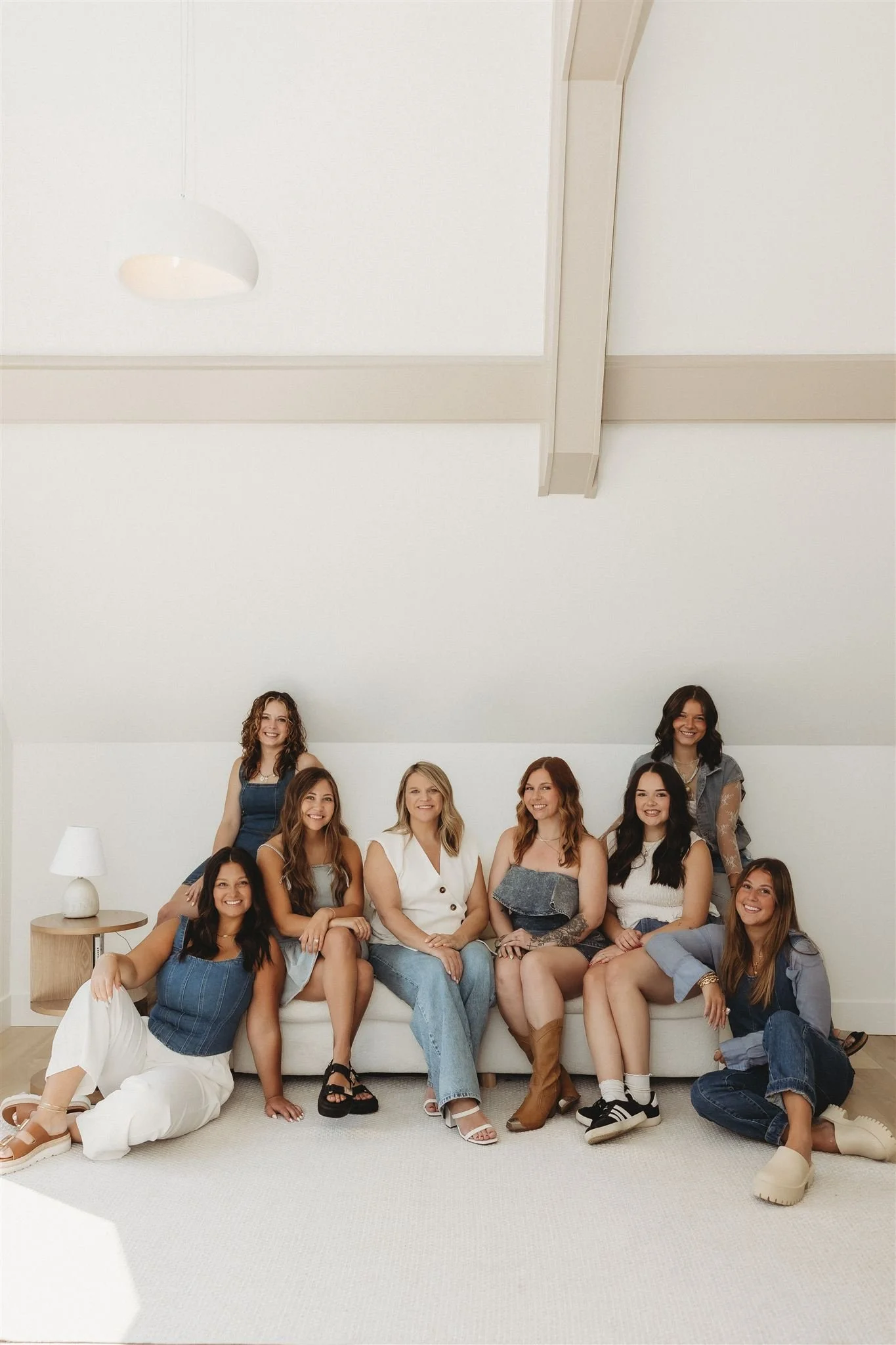 Group of nine women sitting on a white sofa in a bright room with white walls and ceiling. Some women are standing, some are sitting, and they are all smiling.