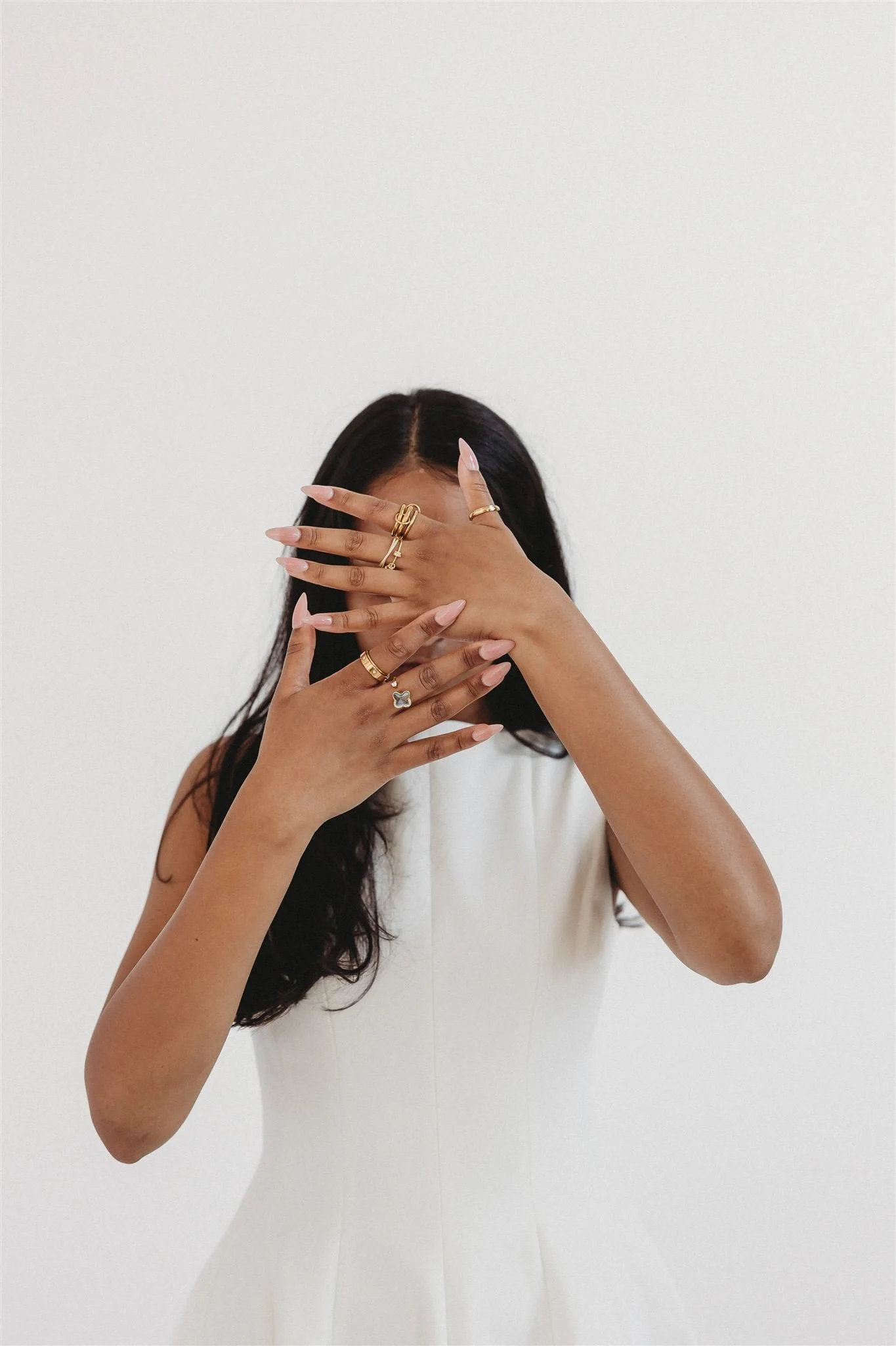 A woman wearing a white sleeveless dress is covering her face with her left hand, showing multiple rings on her fingers against a plain white background.