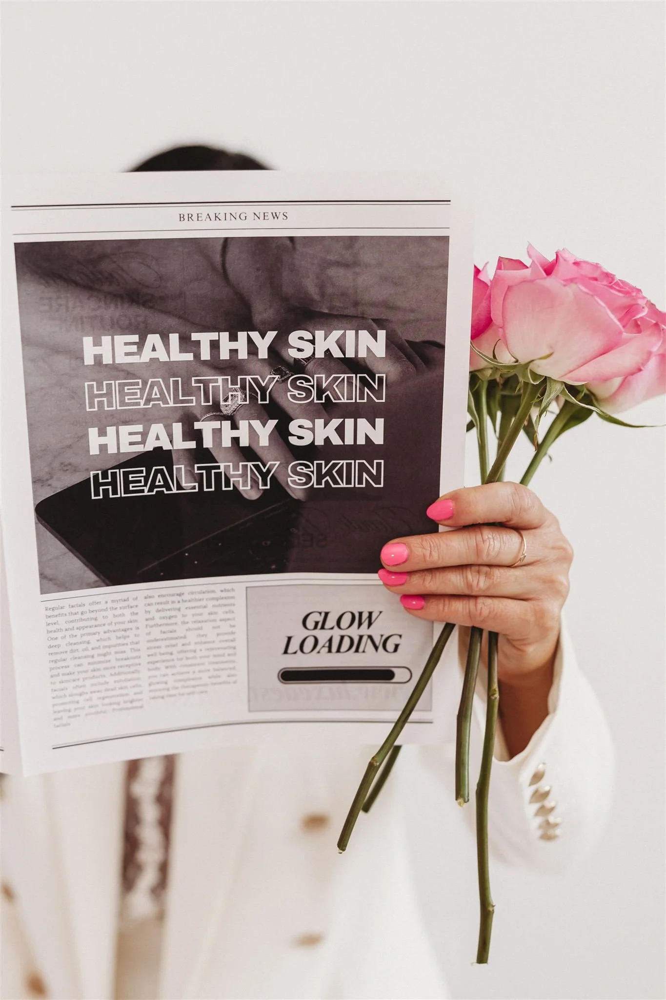 A person holds a newspaper with a headline about healthy skin, partially covering their face, and is holding a small bouquet of pink roses.
