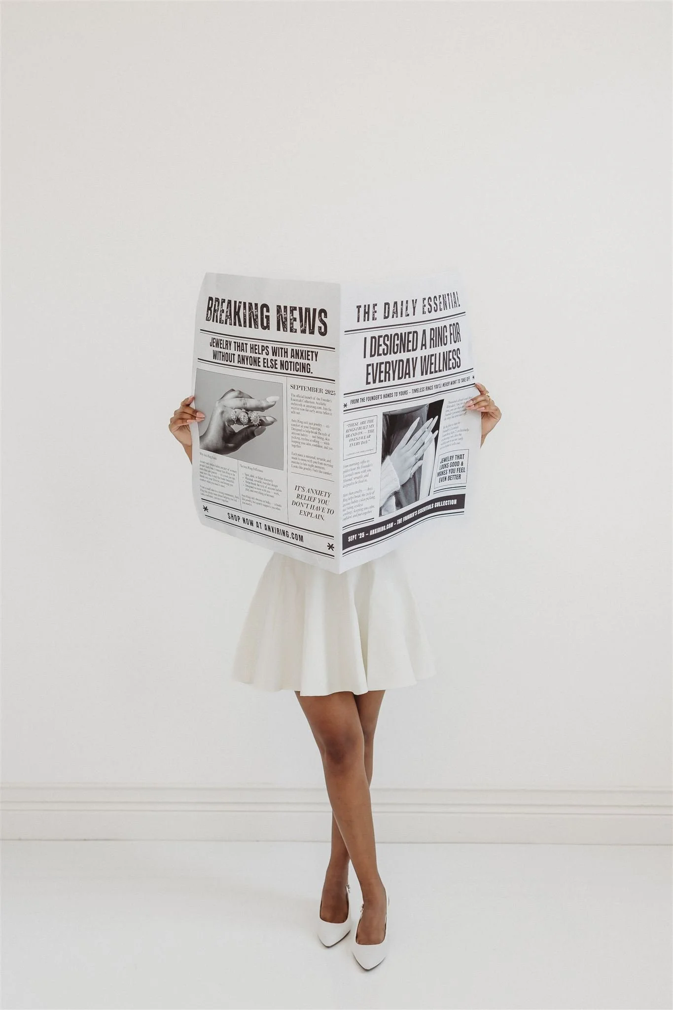 A woman in a white dress and high heels stands against a plain white background, holding and reading a large newspaper that covers her face and upper body.