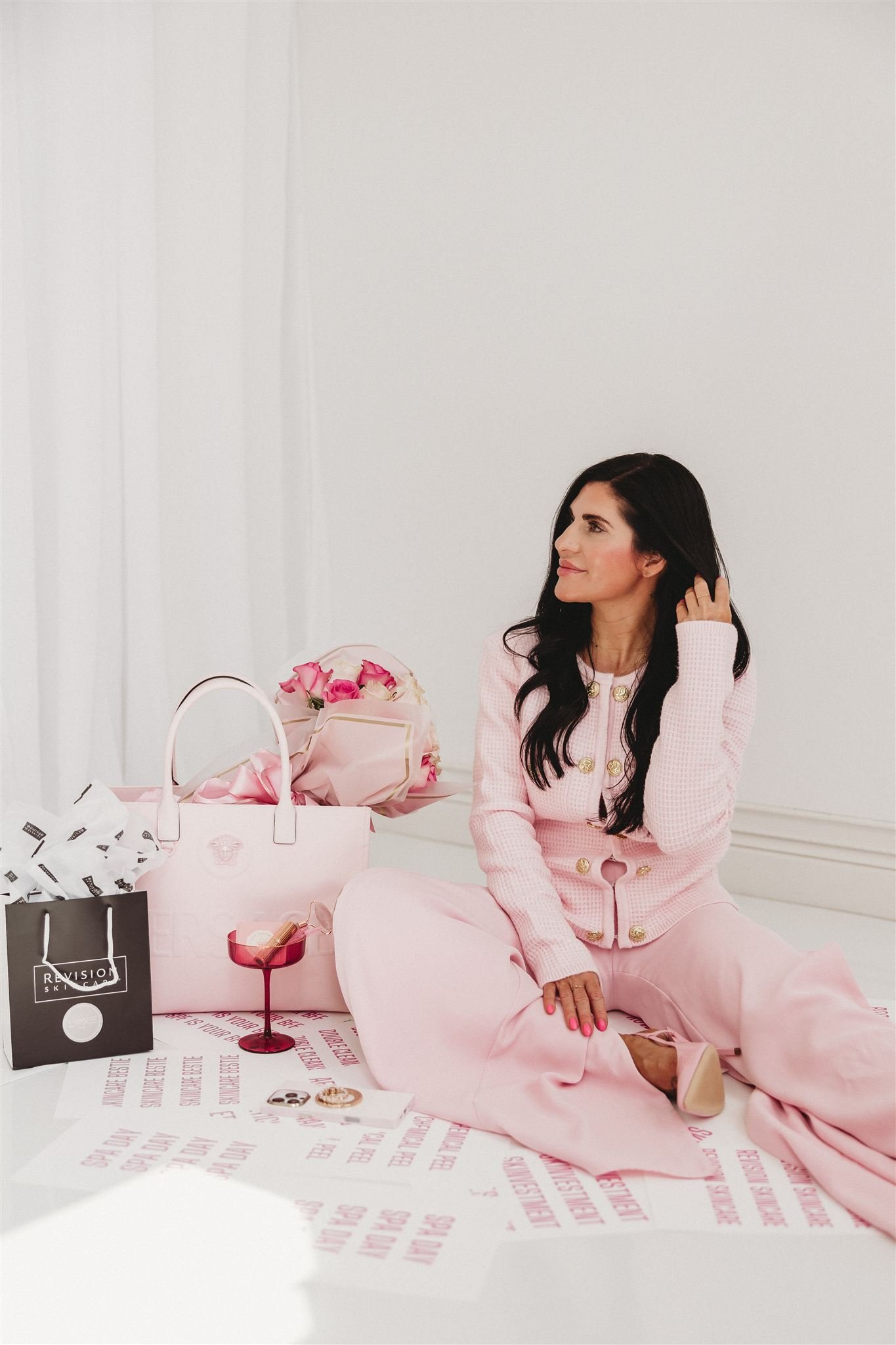 A woman with long dark hair, wearing a pink jacket and matching pants, sitting on the floor next to a table with pink and white flowers, shopping bags, and a red candle holder. The background is plain white.