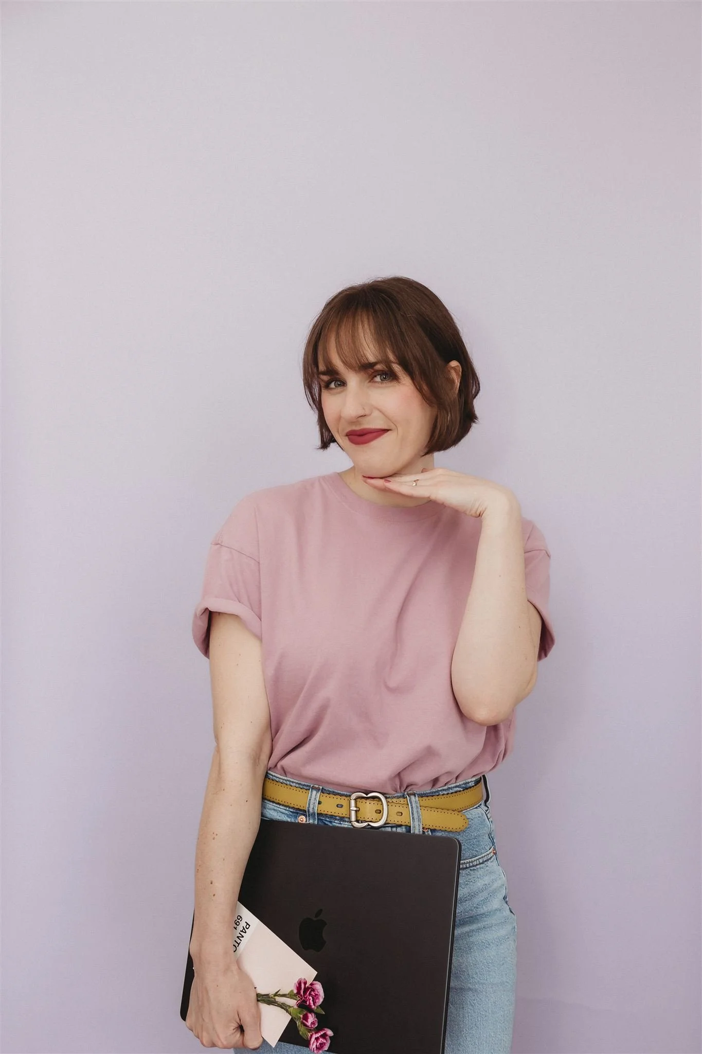 A woman with short brown hair and bangs poses against a plain lilac background. She is wearing a pink t-shirt and light blue jeans with a yellow belt. She is holding a laptop with a black logo and a small bouquet of pink flowers in her left hand. Her right hand is resting on her chin, and she is smiling at the camera.