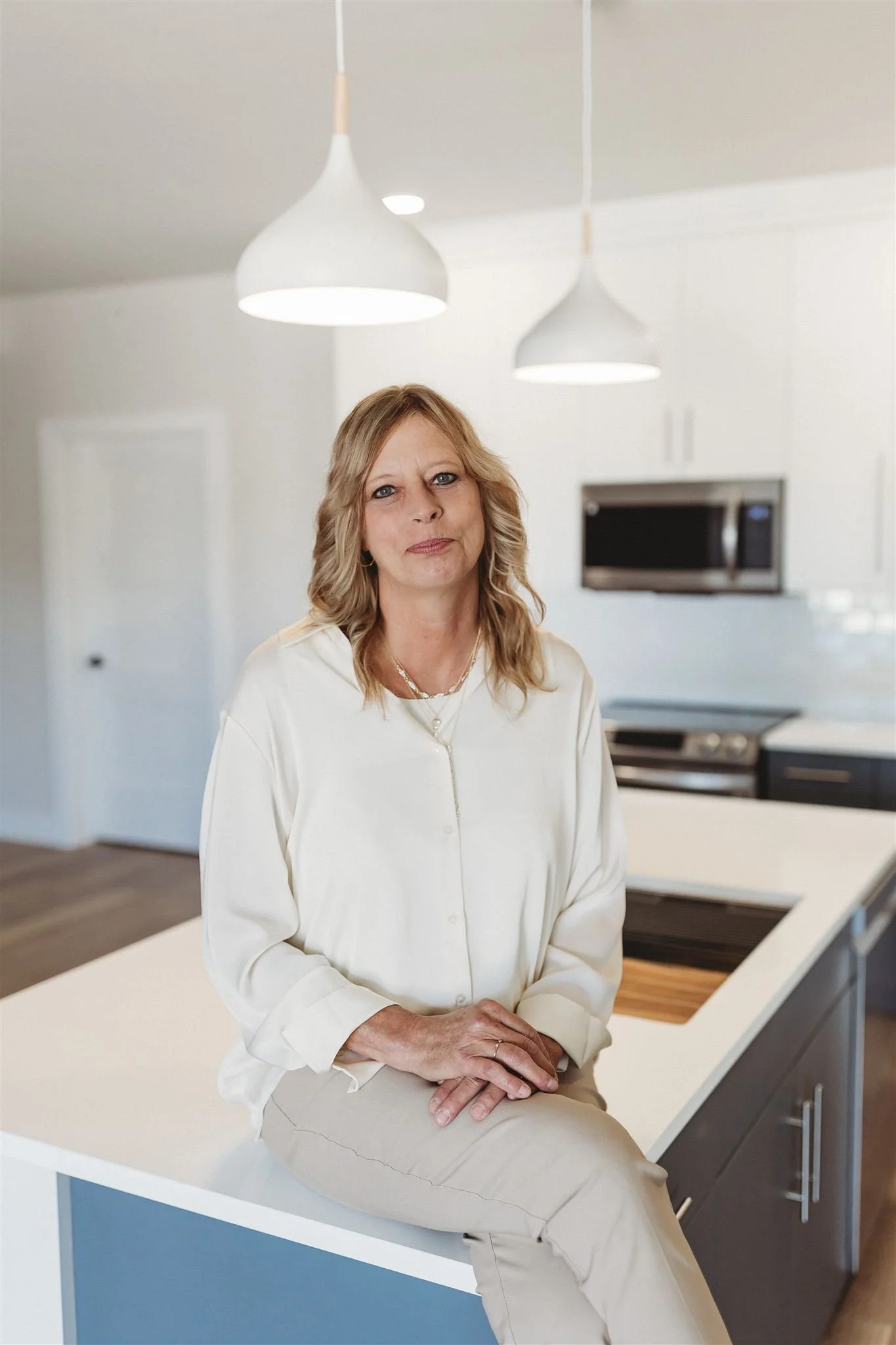 A woman sitting on a kitchen island in a modern kitchen with white cabinets and stainless steel appliances, including a microwave and oven, under two hanging pendant lights.