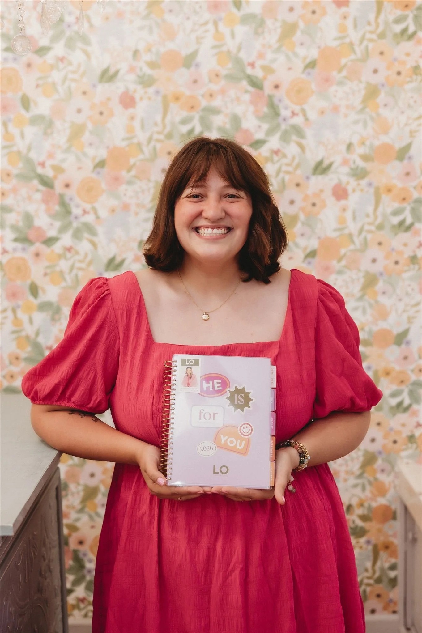 A woman with shoulder-length brown hair, wearing a red dress with puffed short sleeves, smiling and holding a spiral notebook with colorful stickers and tabs, standing in front of a floral-patterned wall.