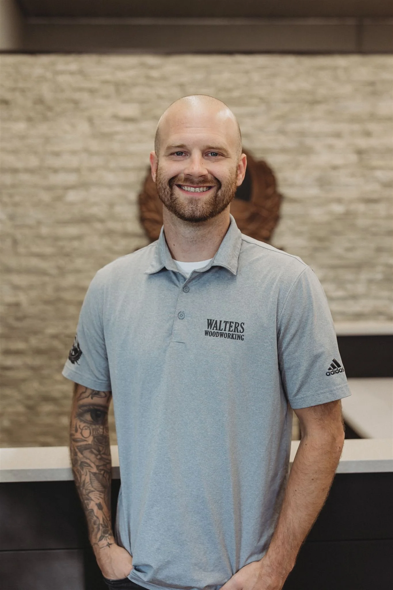 A smiling man with a beard and tattoos on his left arm, wearing a gray polo shirt with 'WALTERS WOODWORKING' logo, standing in front of a textured beige wall.
