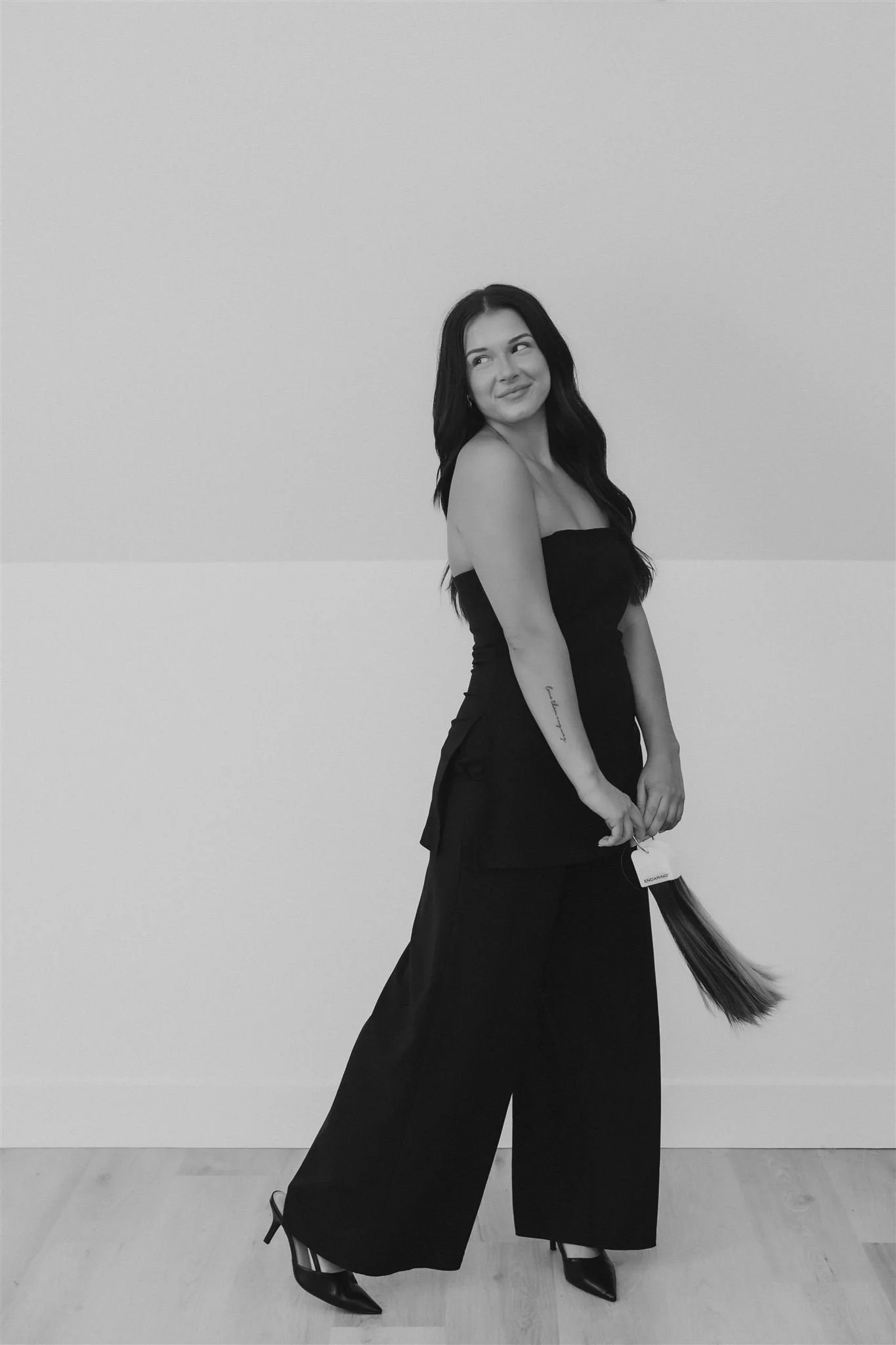 A woman with long dark hair in a strapless black dress holds a bundle of hair extensions. She is smiling and standing against a plain light-colored wall.