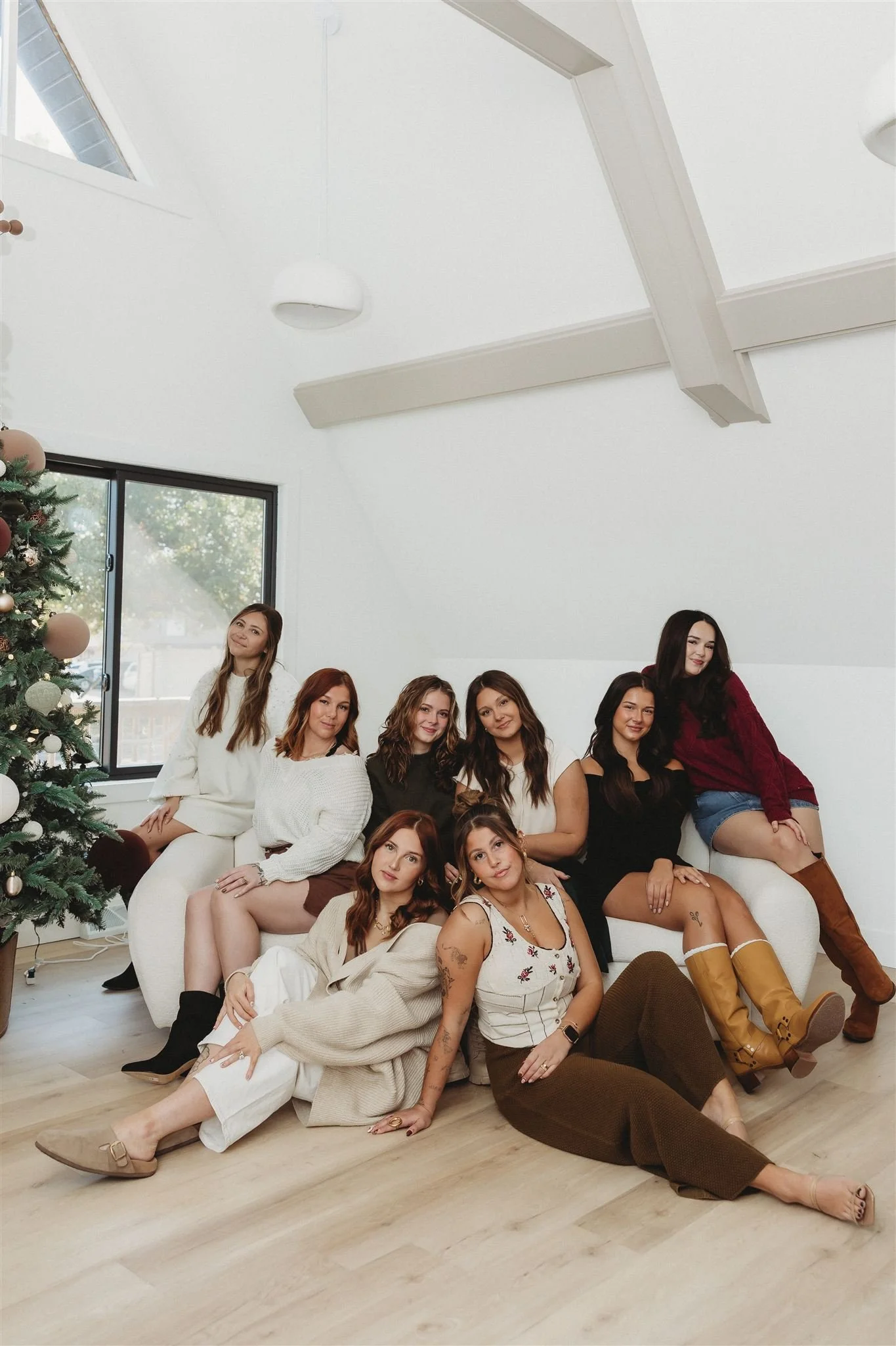 A group of nine women sitting and lying on the floor and a sofa in a bright, white room with a Christmas tree, large window, and vaulted ceiling.