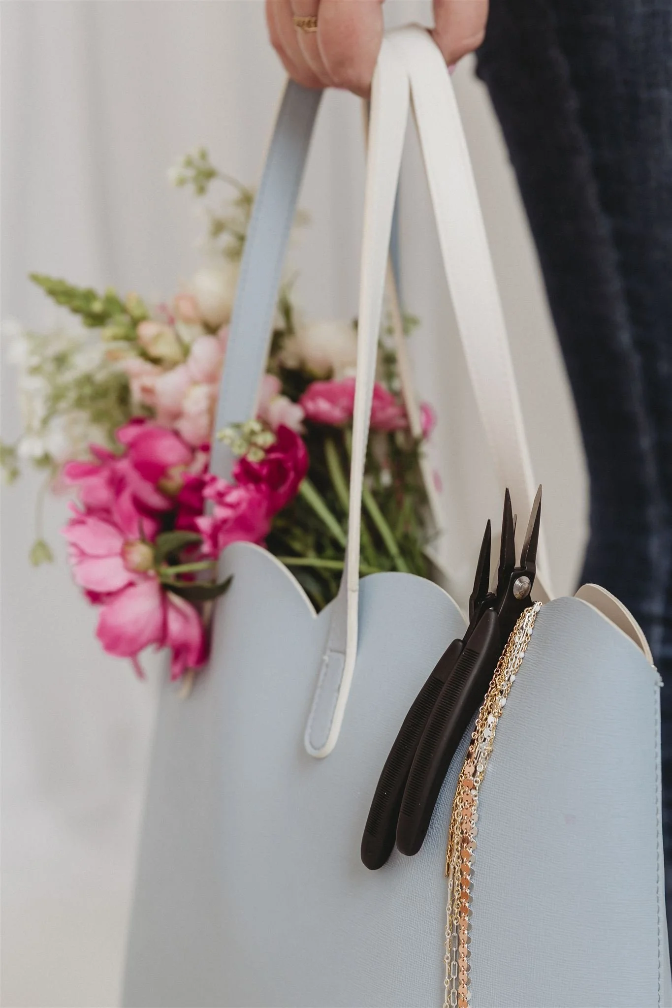 A light blue flower basket hanging from a person’s hand, containing pink and white flowers. A pair of black scissors and some gold jewelry are hanging from the basket.