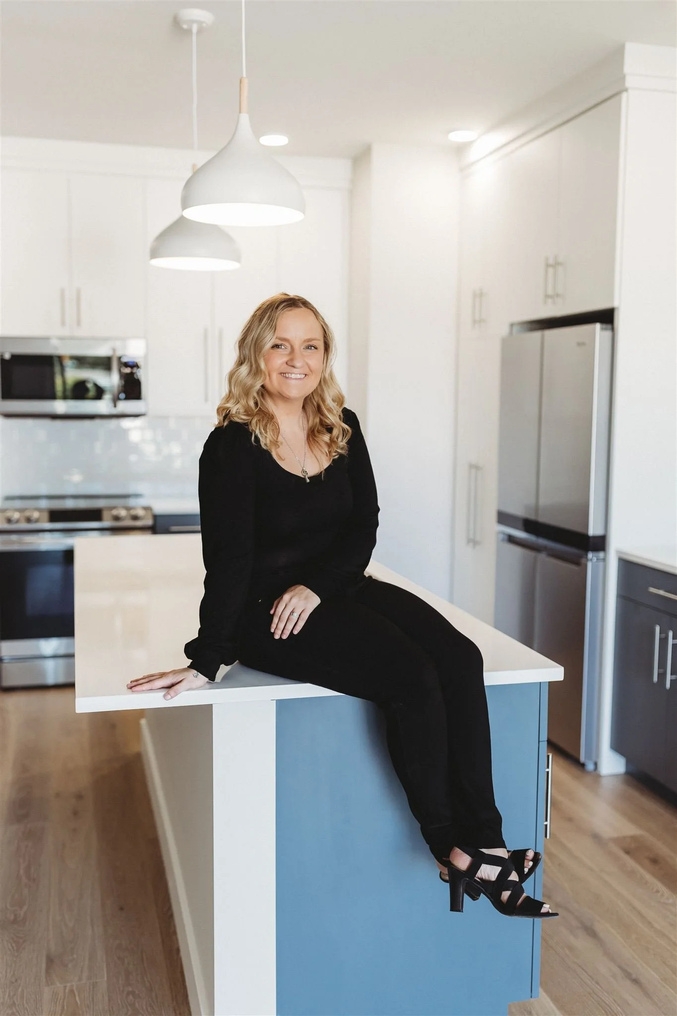 A woman with blonde hair, dressed in black, smiling while sitting on a kitchen island in a modern kitchen with white cabinets and stainless steel appliances.