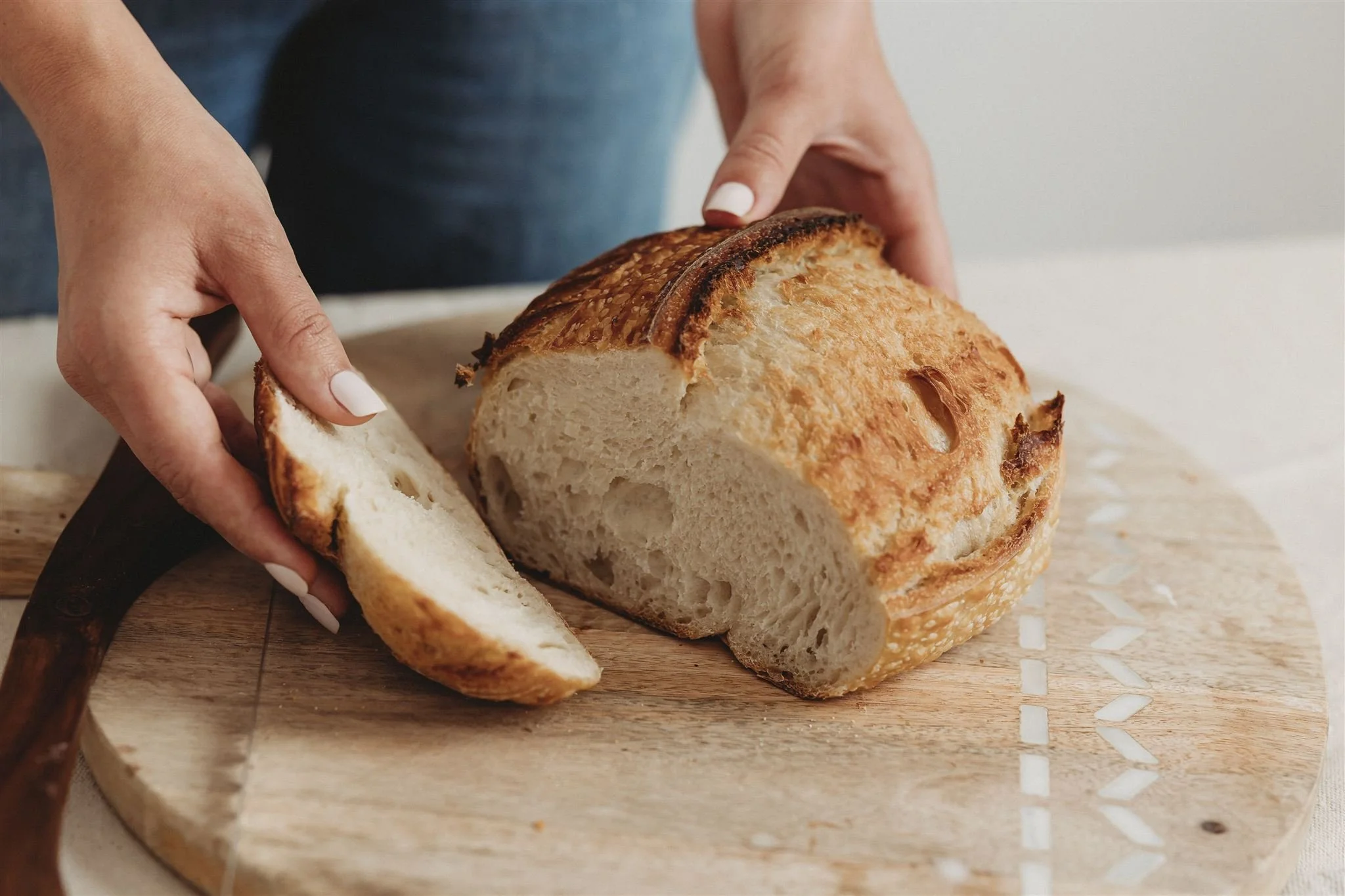 Close-up brand photo of Wild Flour Sourdough Bakehouse baker slicing a rustic artisan sourdough boule on a round wooden cutting board, showing a golden crust and open crumb — Central Illinois brand photography
