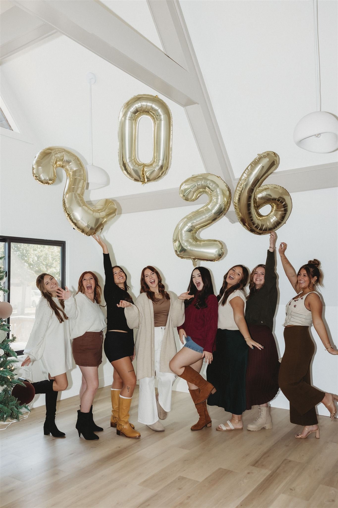 Group of women celebrating in a room with gold balloons spelling '2026', holiday decorations, and large windows.