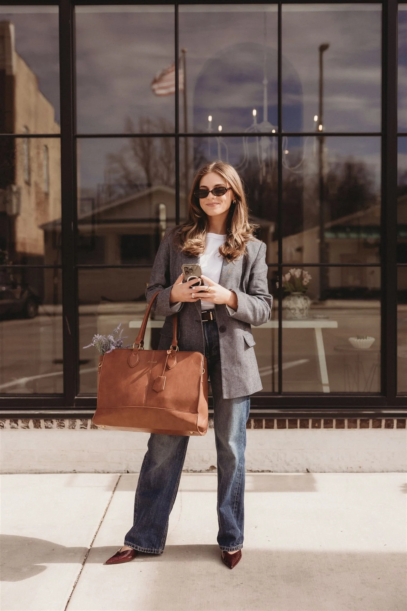 A woman with long brown hair, sunglasses, a gray blazer, white t-shirt, and jeans standing outside in daytime, holding a smartphone and a large brown handbag. There are reflections of buildings and an American flag in the window behind her.