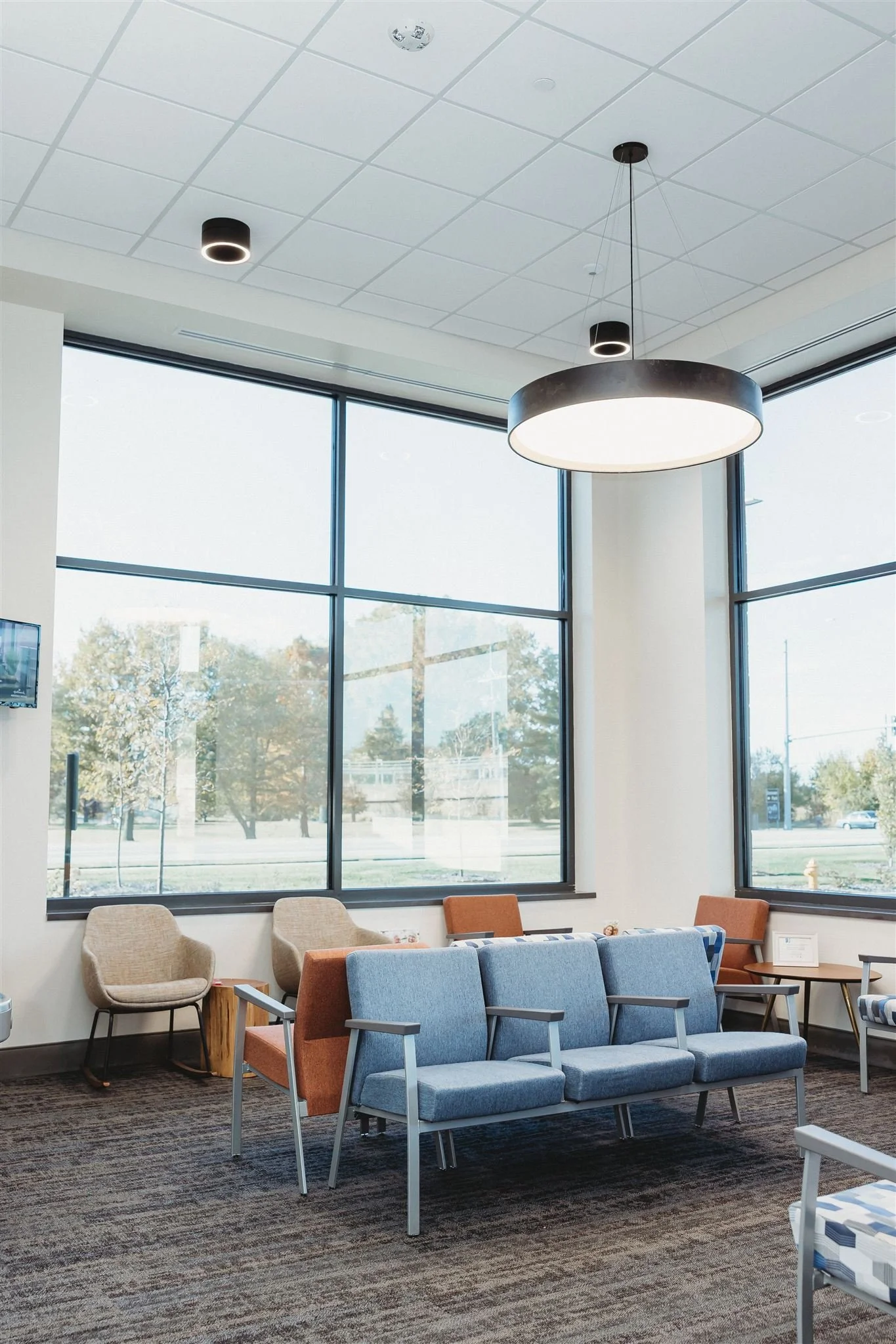 A waiting area with blue and beige chairs, a small wooden table, and large floor-to-ceiling windows with an outside view of trees and a street.