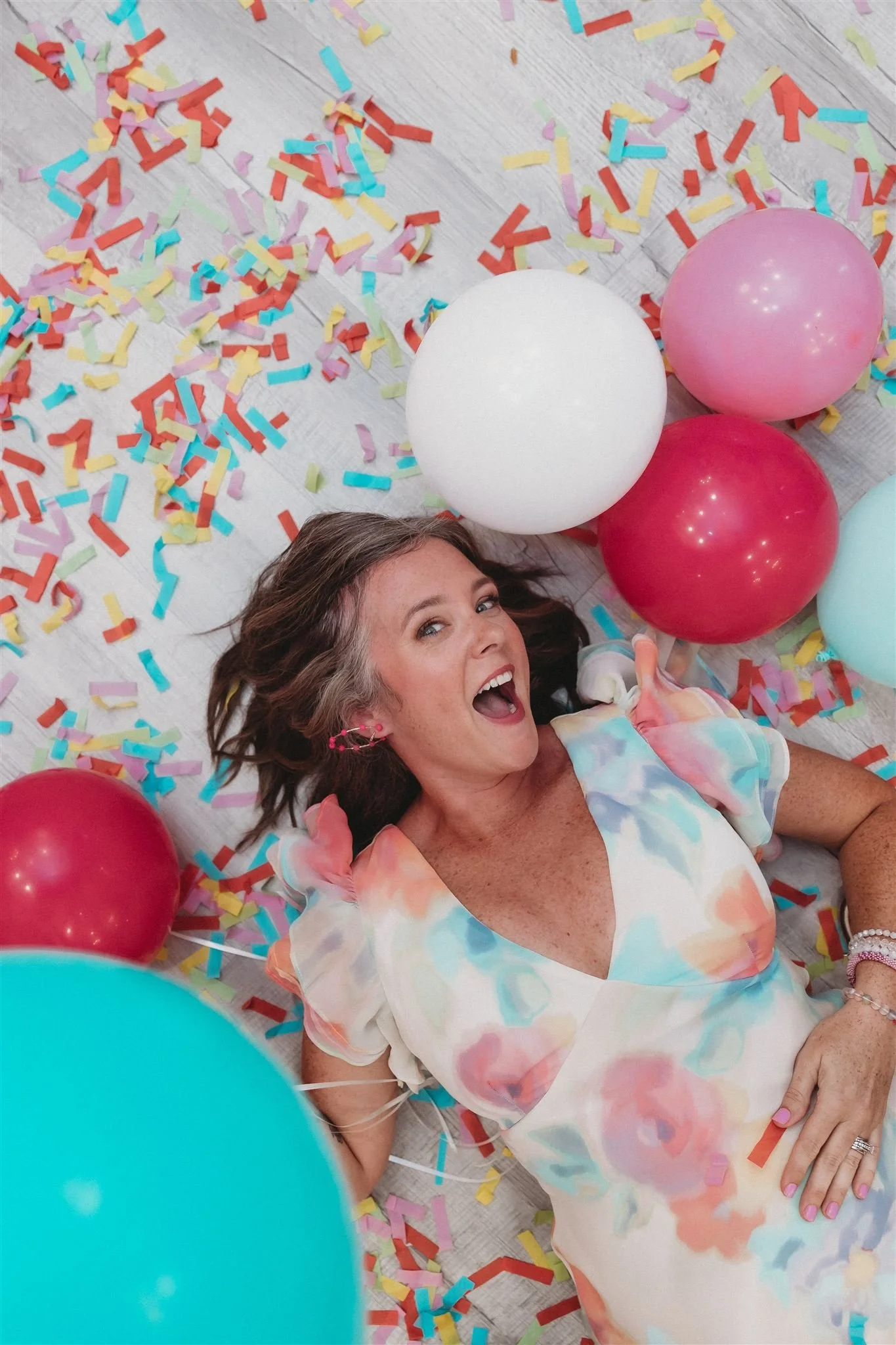A woman lying on the floor surrounded by colorful confetti and balloons, smiling and looking up at the camera.