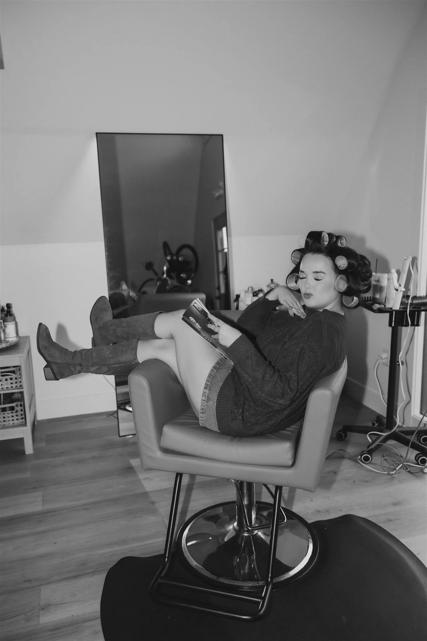 A woman with hair rollers sitting in a salon chair, reading a magazine.
