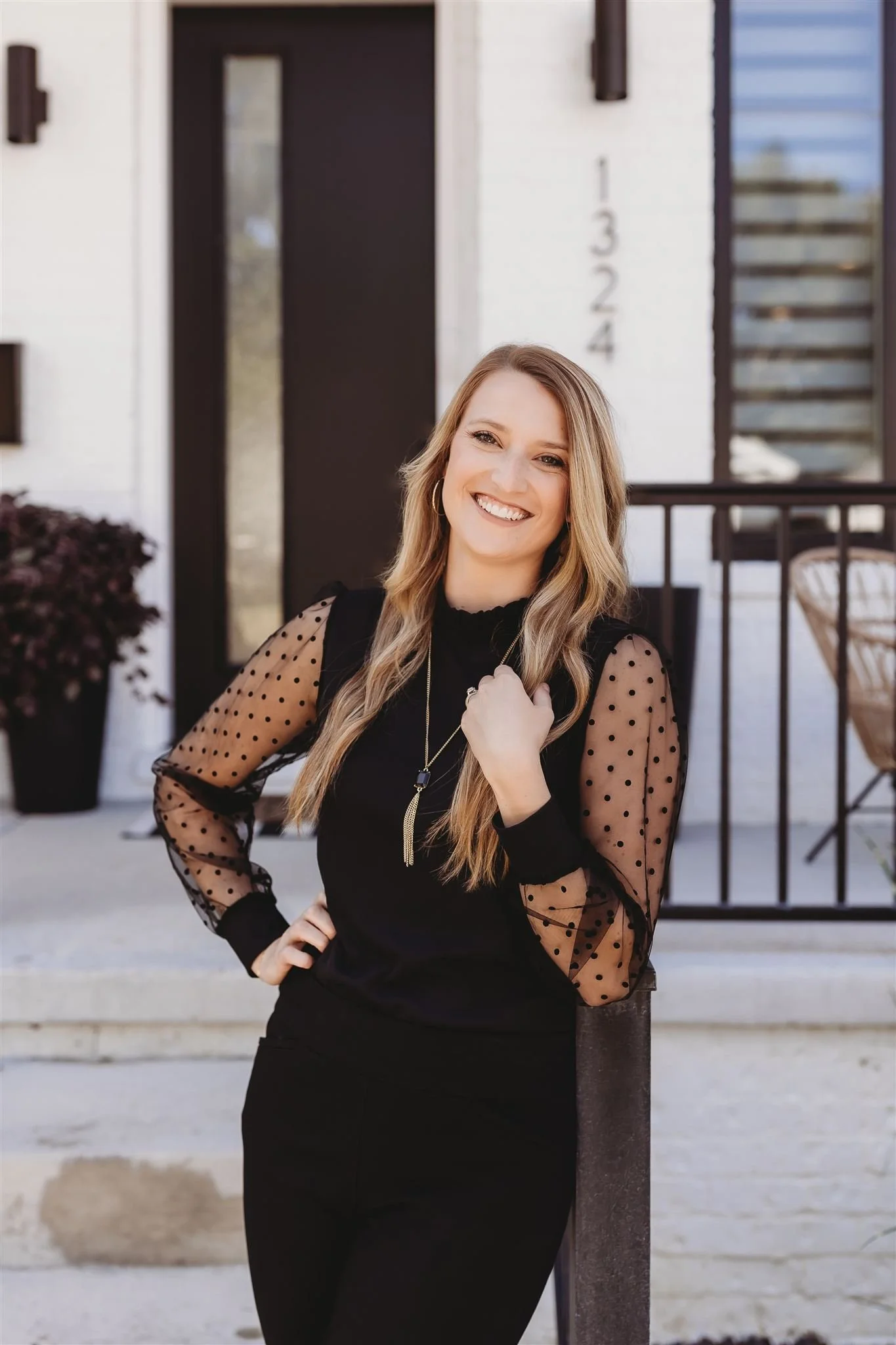 A woman with long, wavy blonde hair smiling and standing outside in front of a modern house with a black door and white exterior. She is wearing a black top with sheer, polka-dot sleeves and dark pants.