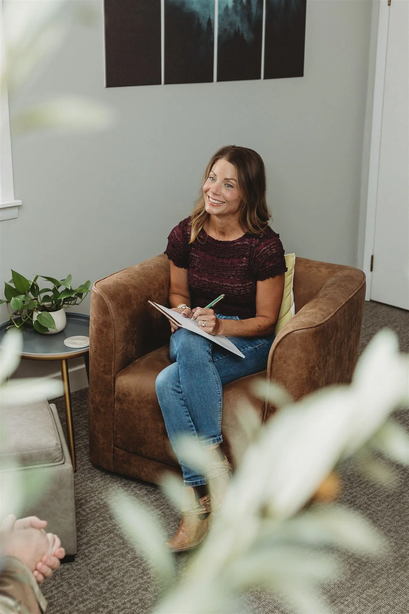 A woman sitting in a brown armchair, smiling, taking notes with a pen on a notepad in a cozy, well-lit room decorated with plants and artwork.