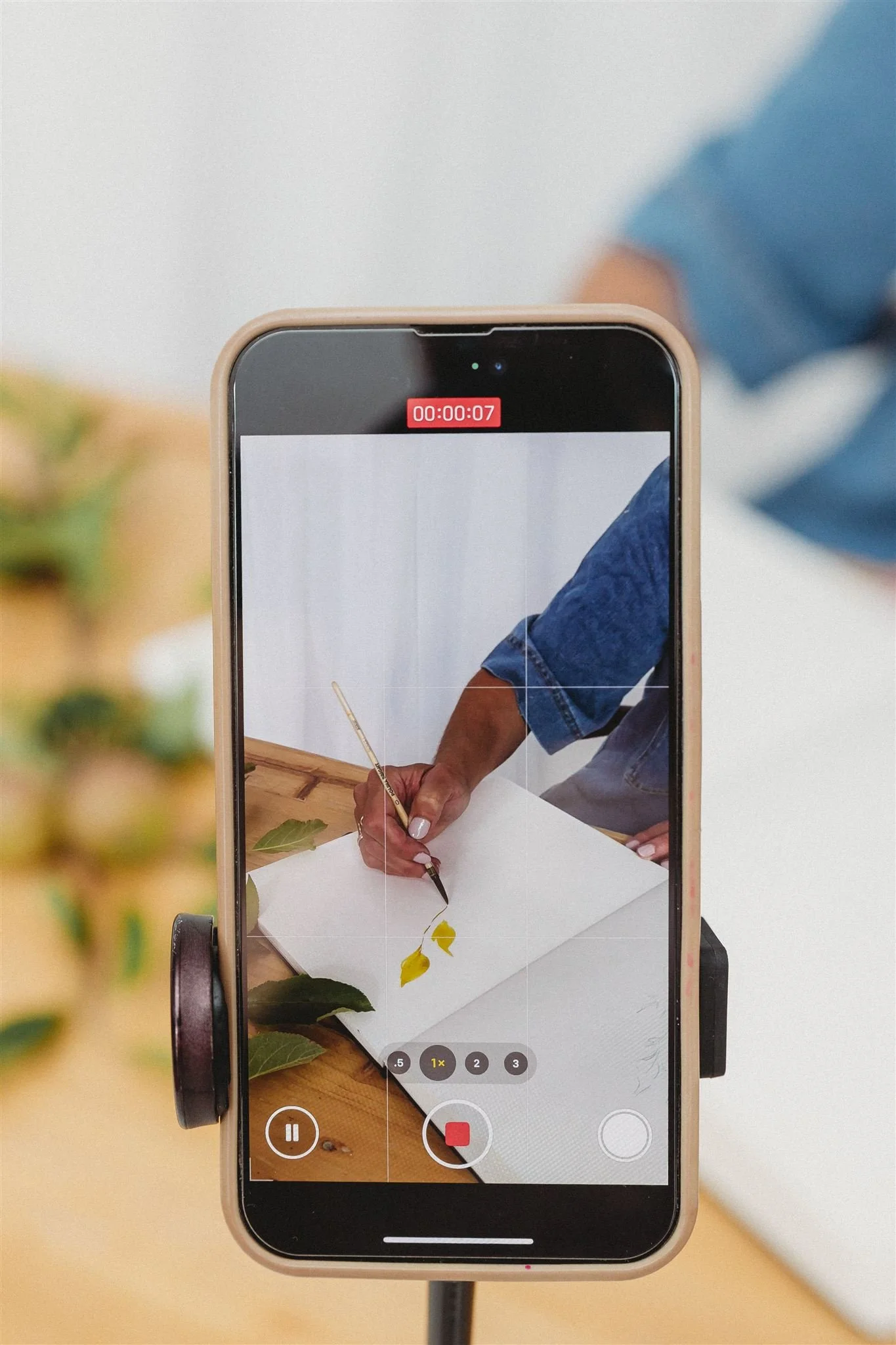 Smartphone mounted on a stand recording a person painting with a brush on white paper on a wooden table.