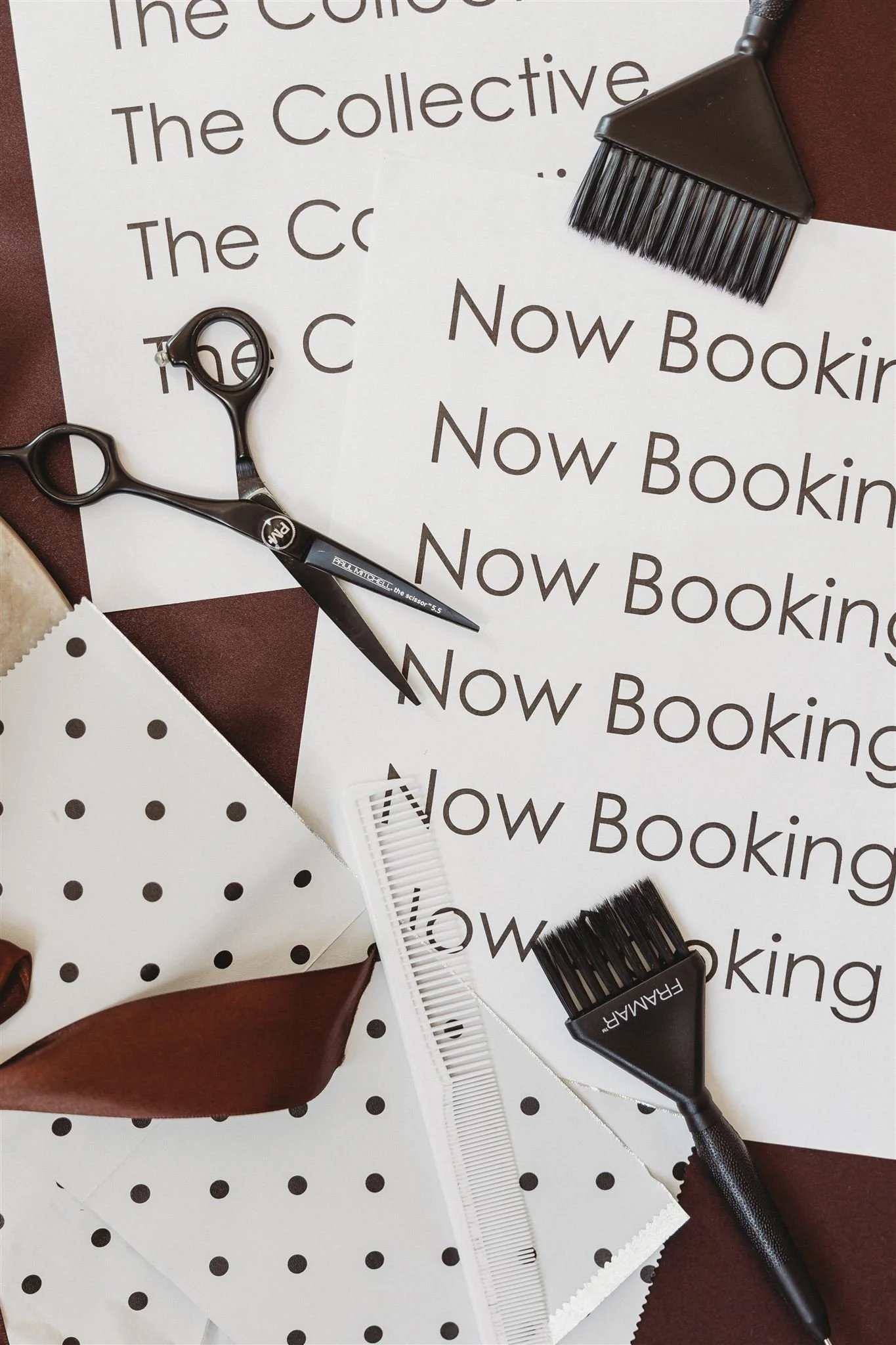 A flat lay of hairdressing tools and printed signs with the words 'Now Booking' on some, polka dot paper, a wide-tooth comb, scissors, and hairbrushes on a polka dot and dark background.