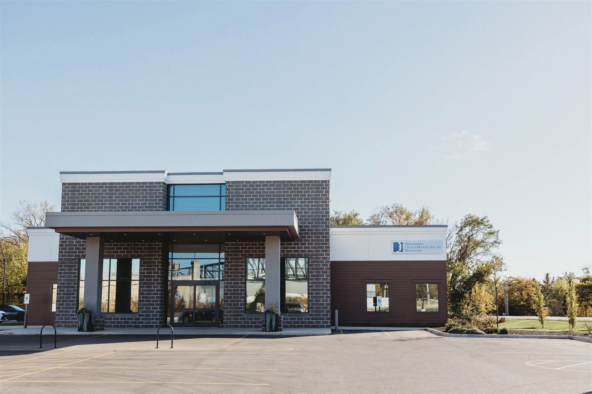 A modern medical office building with a brick and wood facade, large glass entrance, and a parking lot in front, signage indicating it houses an oral and maxillofacial surgery practice.